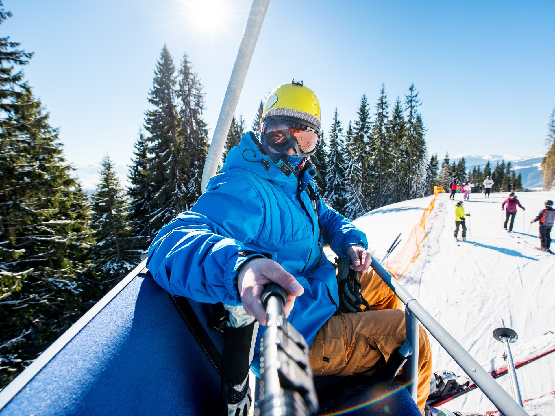 Eine Person auf einem Skilift, die einen Selfie-Stick hält, mit Skifahrern auf einer schneebedeckten Piste.