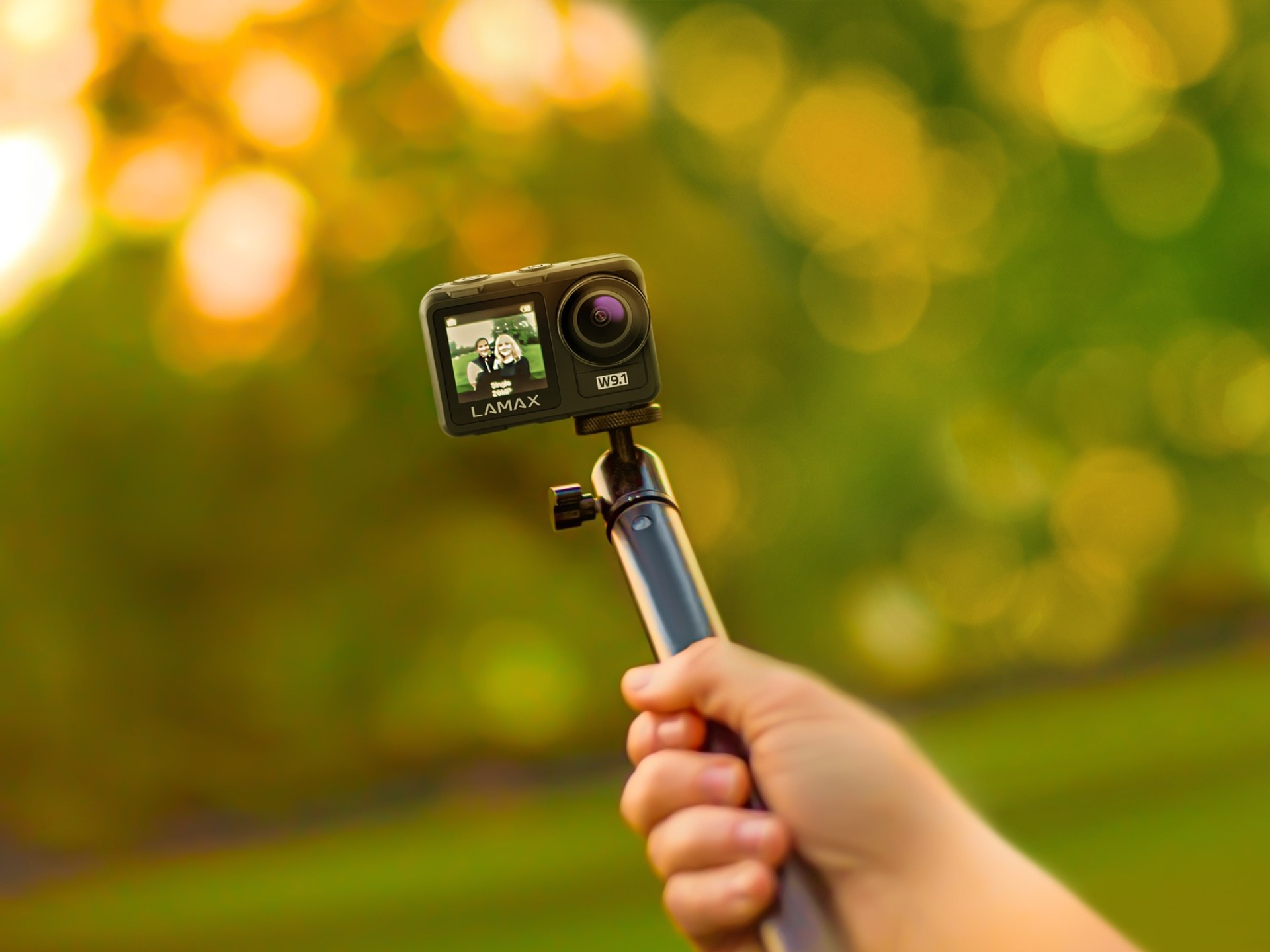 Eine Person hält einen Selfie-Stick mit Kamera, grüner Hintergrund, Bokeh-Effekt.