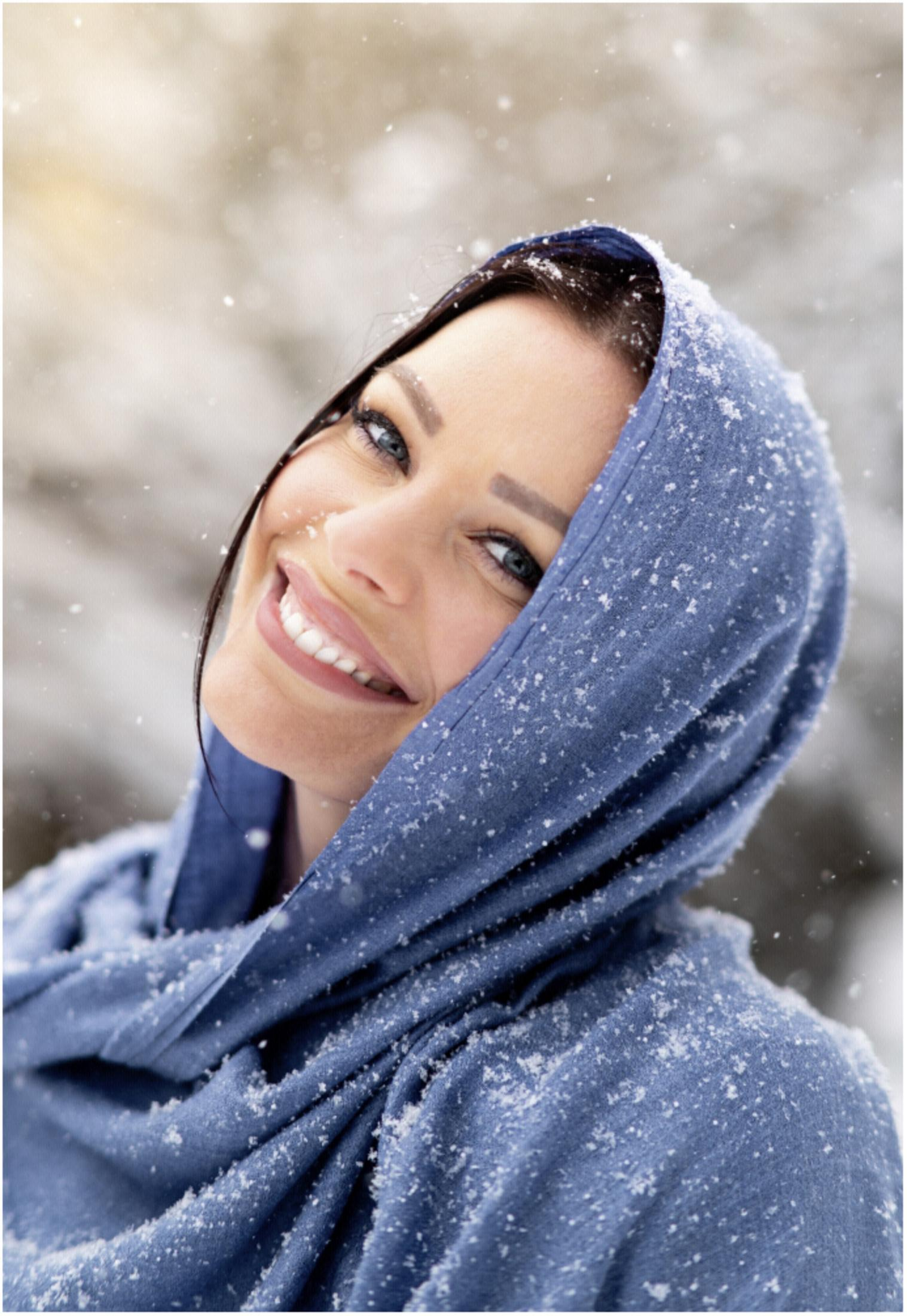 Uśmiechnięta kobieta z niebieskim szalikiem w śnieżnym otoczeniu. Płatki śniegu są widoczne.