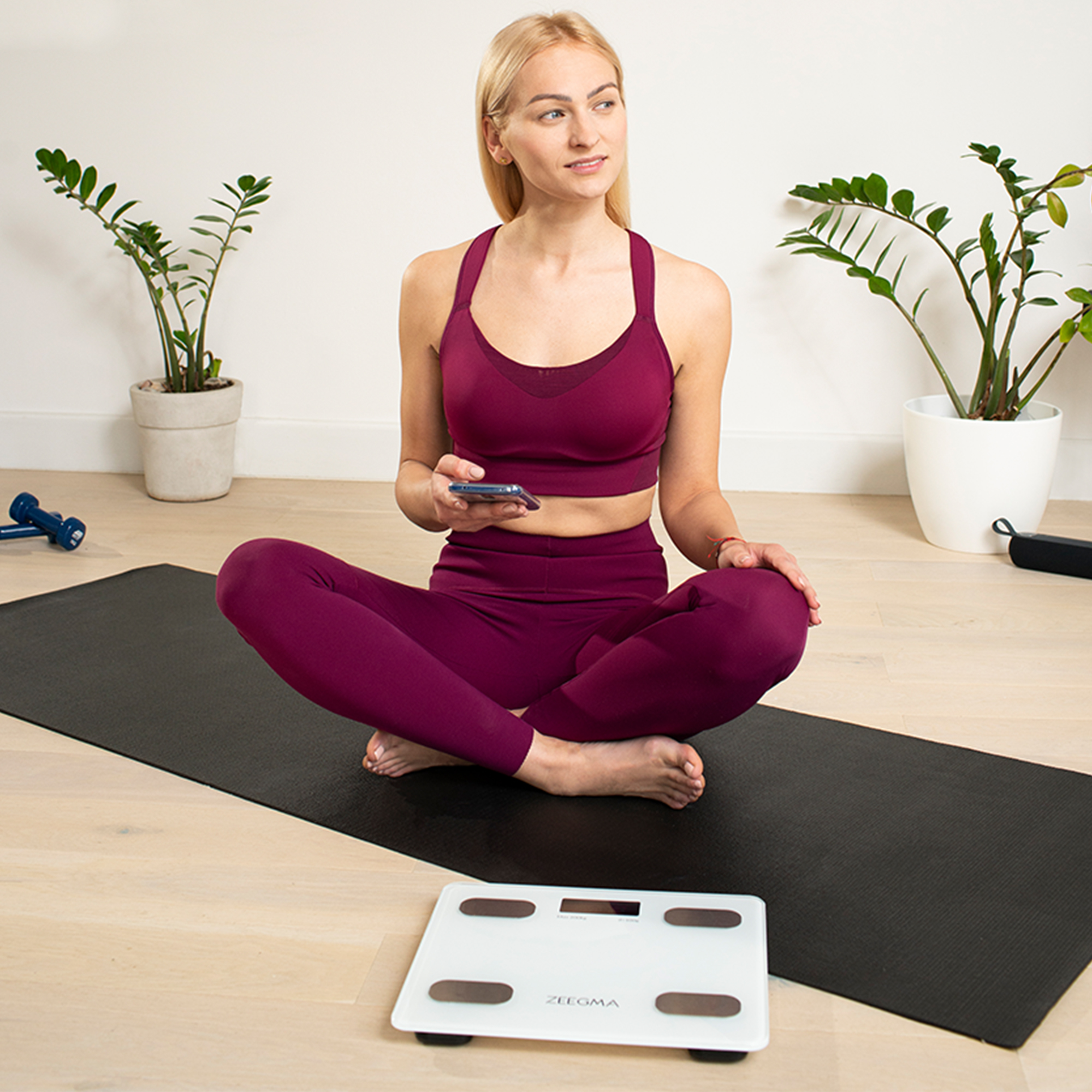 Kobieta w bordowej odzieży sportowej siedzi na czarnej macie, sprawdzając telefon. Widoczna waga i rośliny doniczkowe.