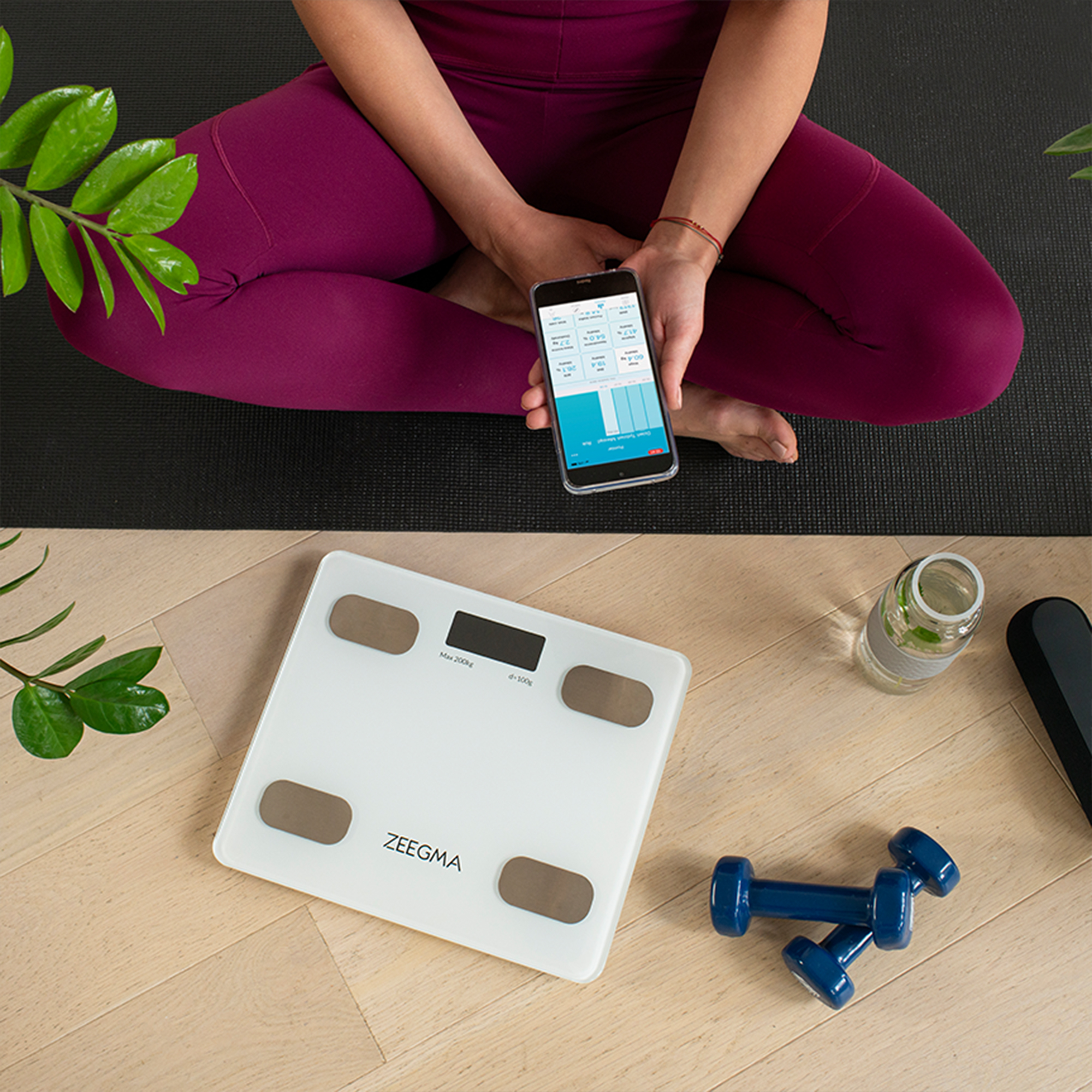 Osoba siedząca ze skrzyżowanymi nogami trzymająca telefon z aplikacją zdrowotną, w pobliżu wagi i hantli.
