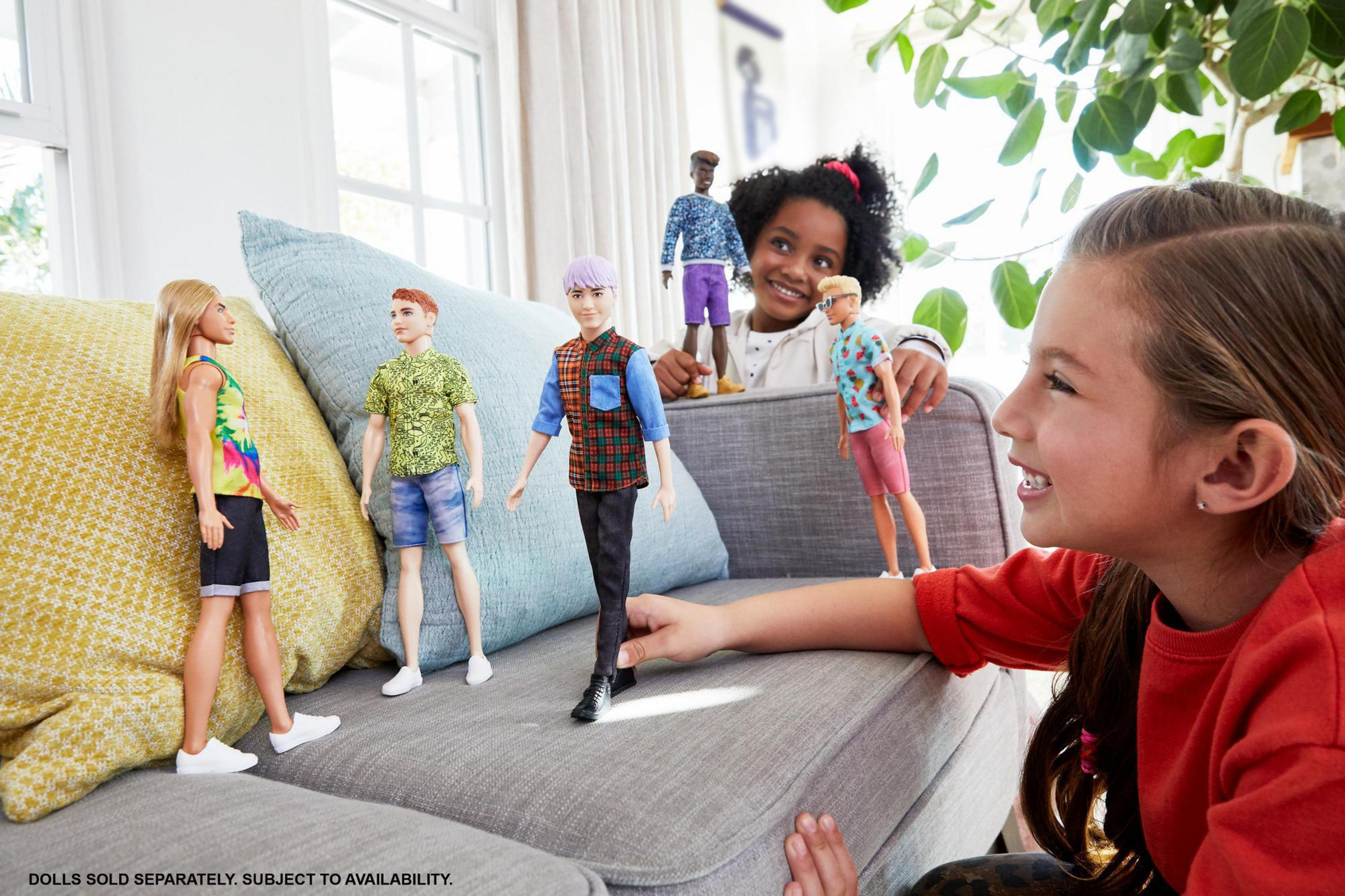 Kinder mit Barbie-Puppen. Ein Mädchen lächelt. Puppen in verschiedenen Outfits auf einer Couch.
