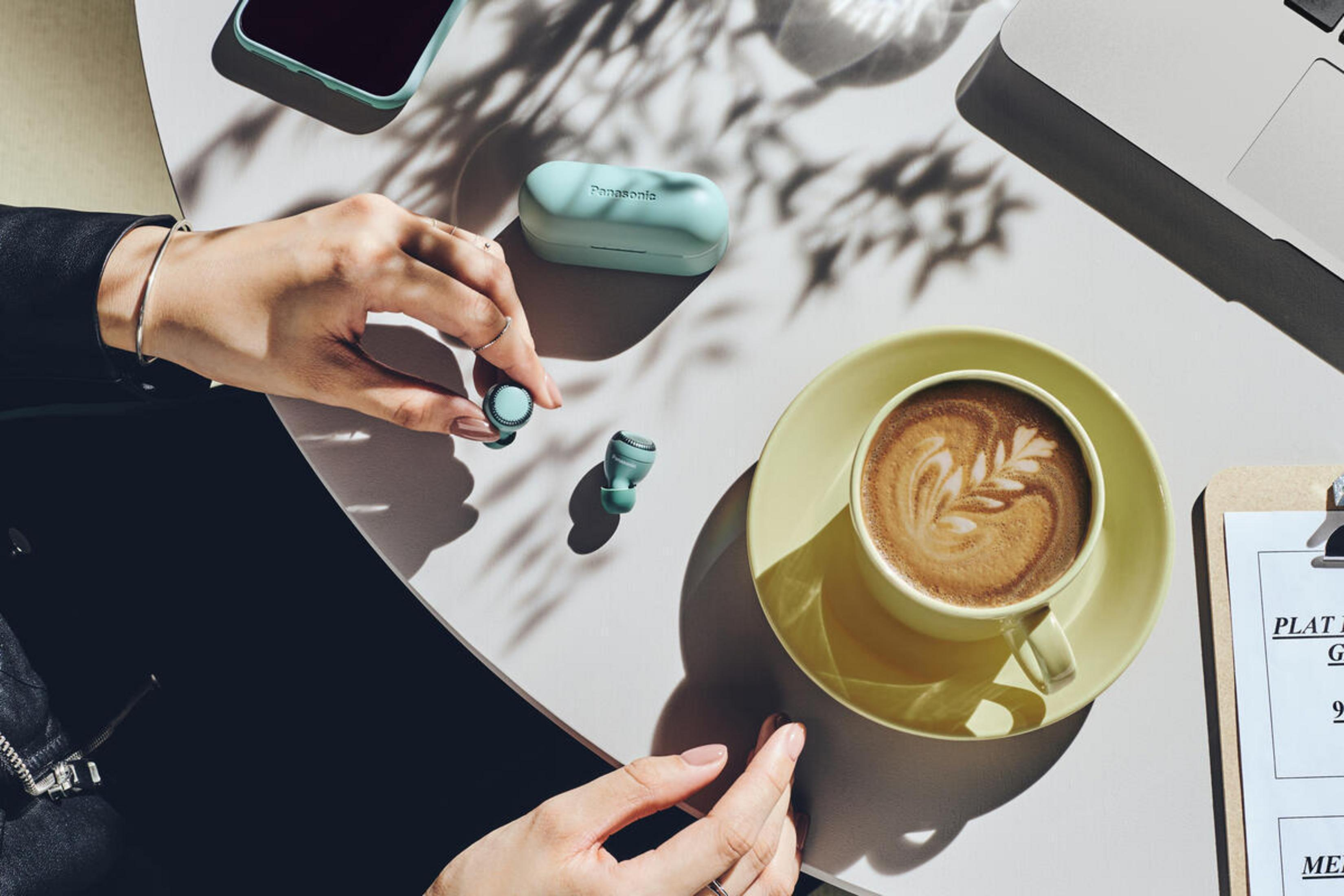 Manos con auriculares, café, funda y teléfono en una mesa.