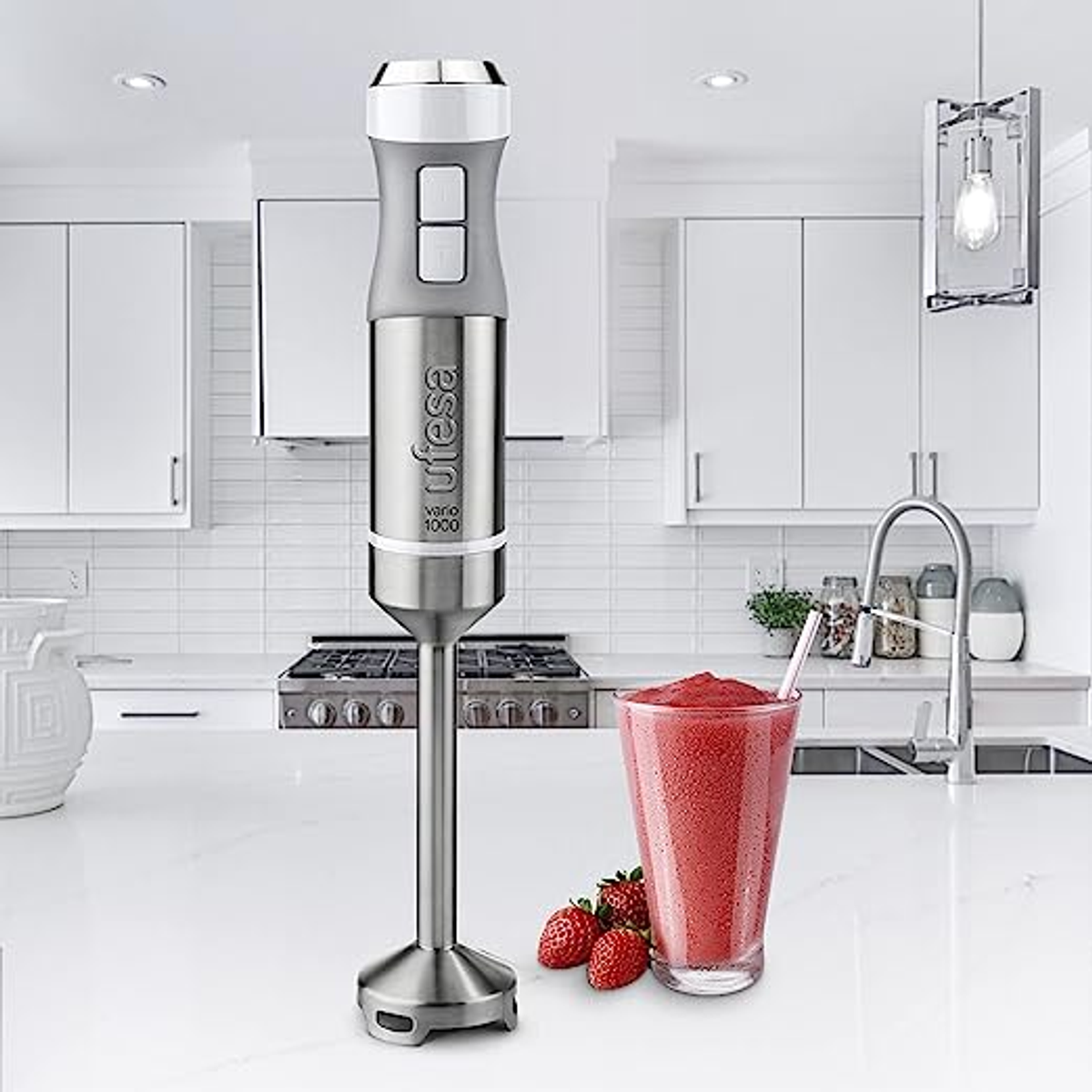 Ein silberner und grauer Handmixer steht neben einem Glas rotem Smoothie und Erdbeeren auf einer weißen Arbeitsplatte.