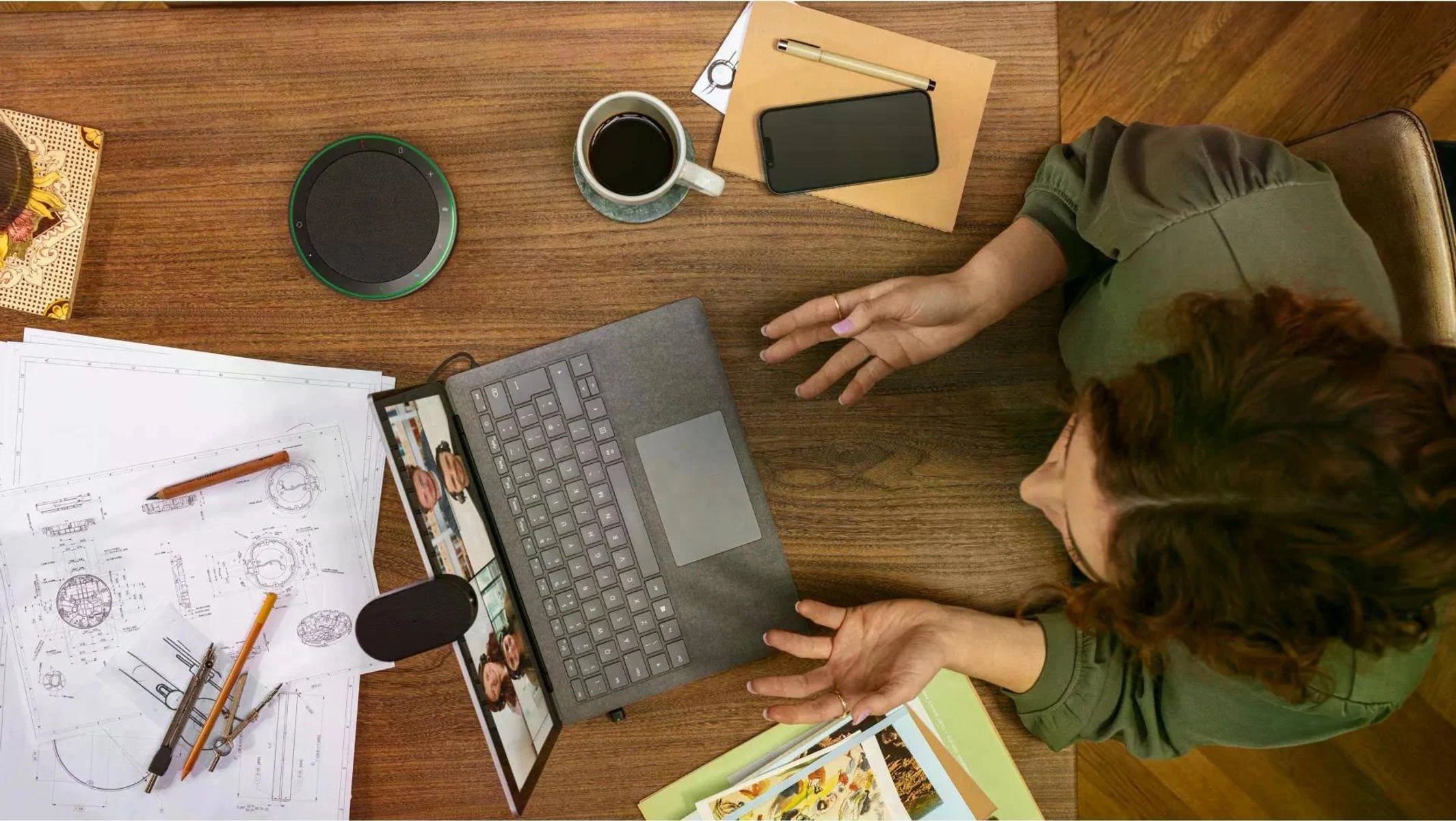 Una persona a un tavolo di legno con laptop, caffè, telefono e progetti.