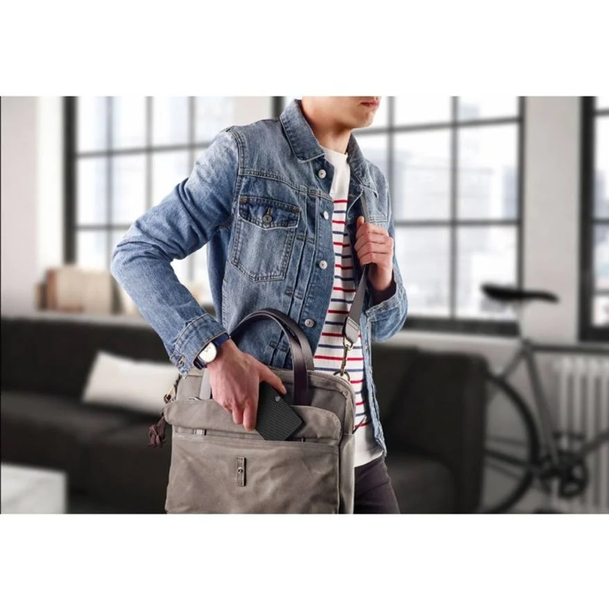 Mężczyzna w dżinsowej kurtce, niosący szarą torbę i czarne urządzenie.