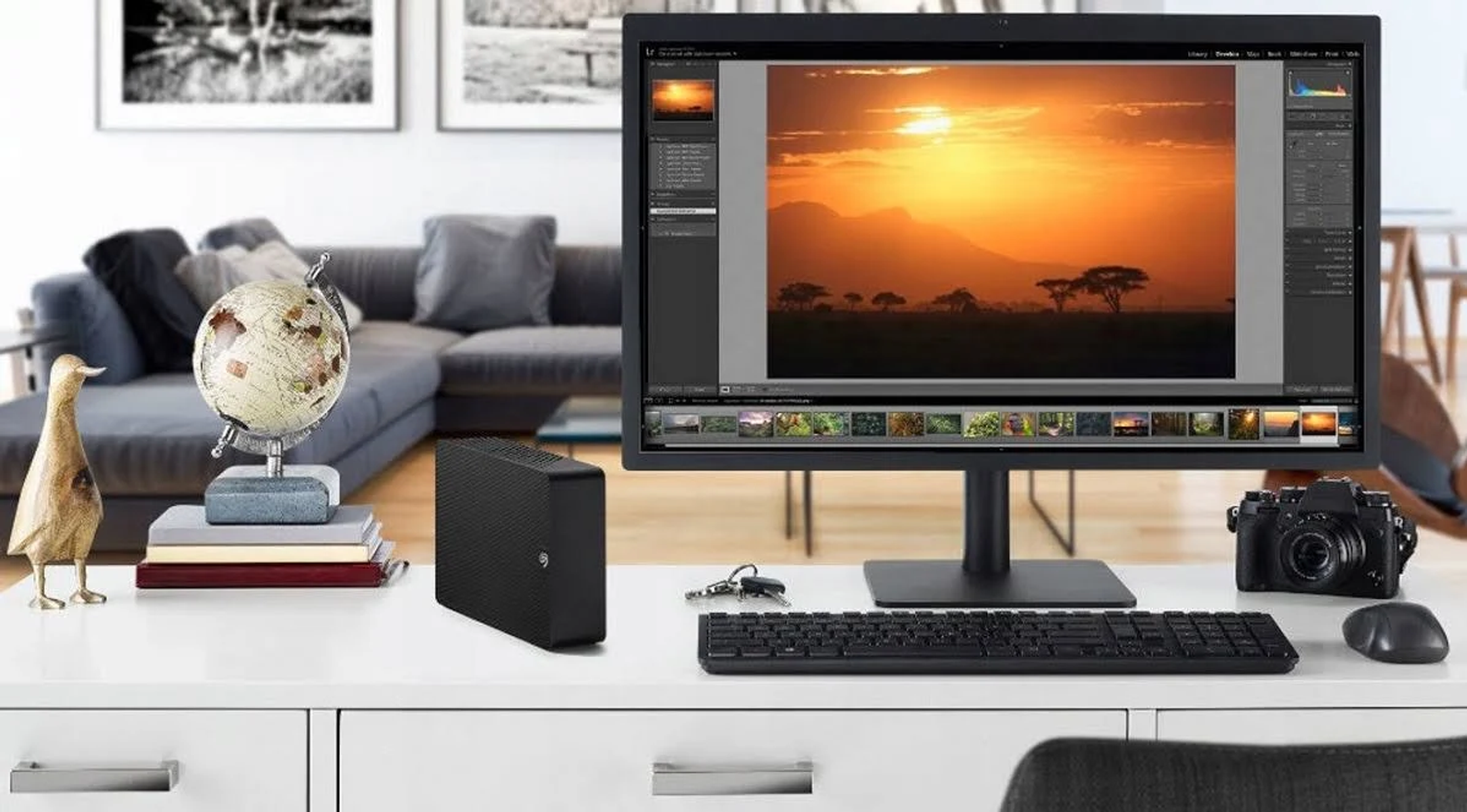 Biurko z monitorem z obrazem zachodu słońca. Klawiatura, aparat i mysz na biurku. Globus i dekoracja z boku.