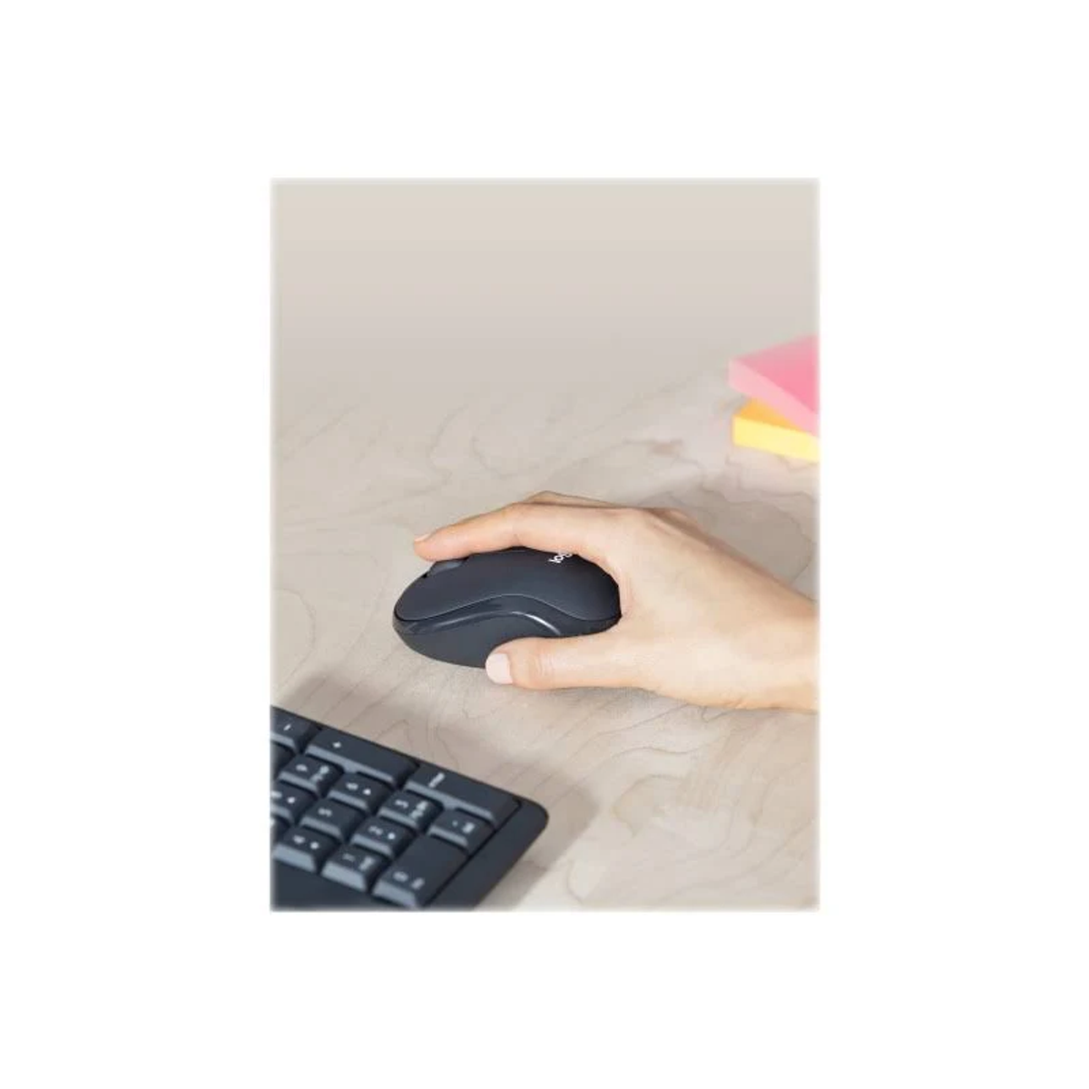 Hand hält eine schwarze Maus auf einem hölzernen Schreibtisch. Tastatur und Haftnotizen sind ebenfalls vorhanden.