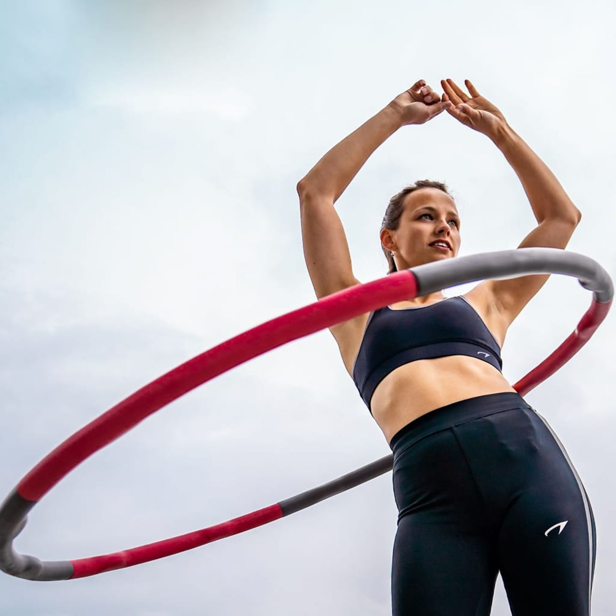Frau mit Hula-Hoop, Arme erhoben. Himmelshintergrund. Trägt schwarze Trainingskleidung.