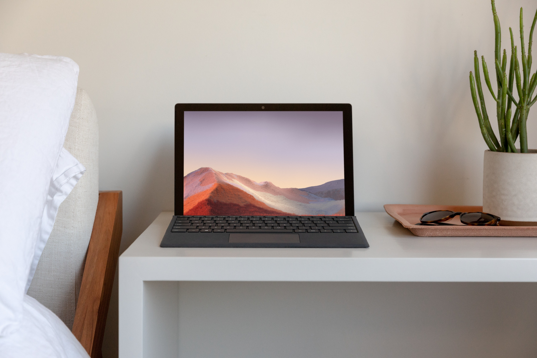 Laptop su un tavolo bianco. Sfondo: letto, comodino, occhiali da sole e pianta.