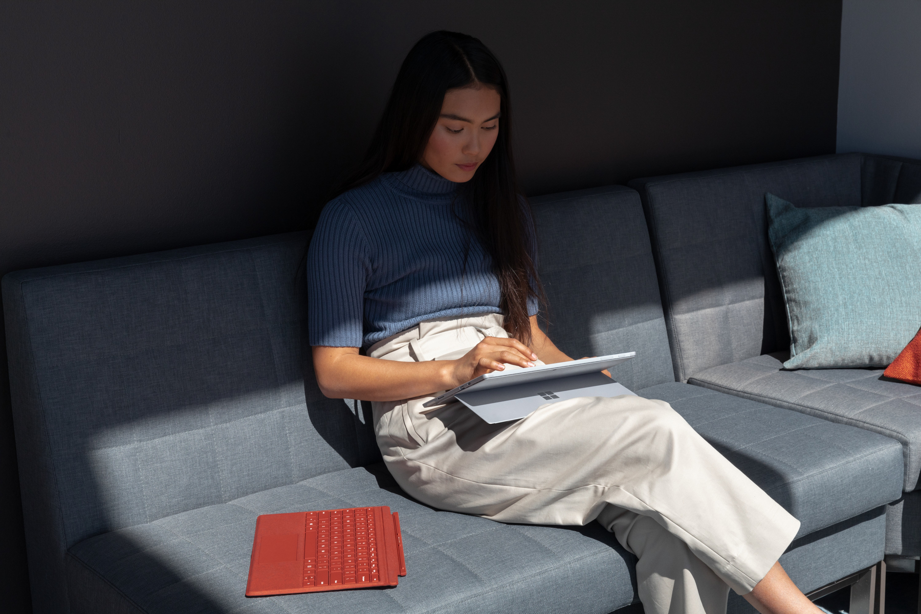 Donna che usa un tablet su un divano grigio. Una tastiera rossa è sul divano. Parete grigio scuro.