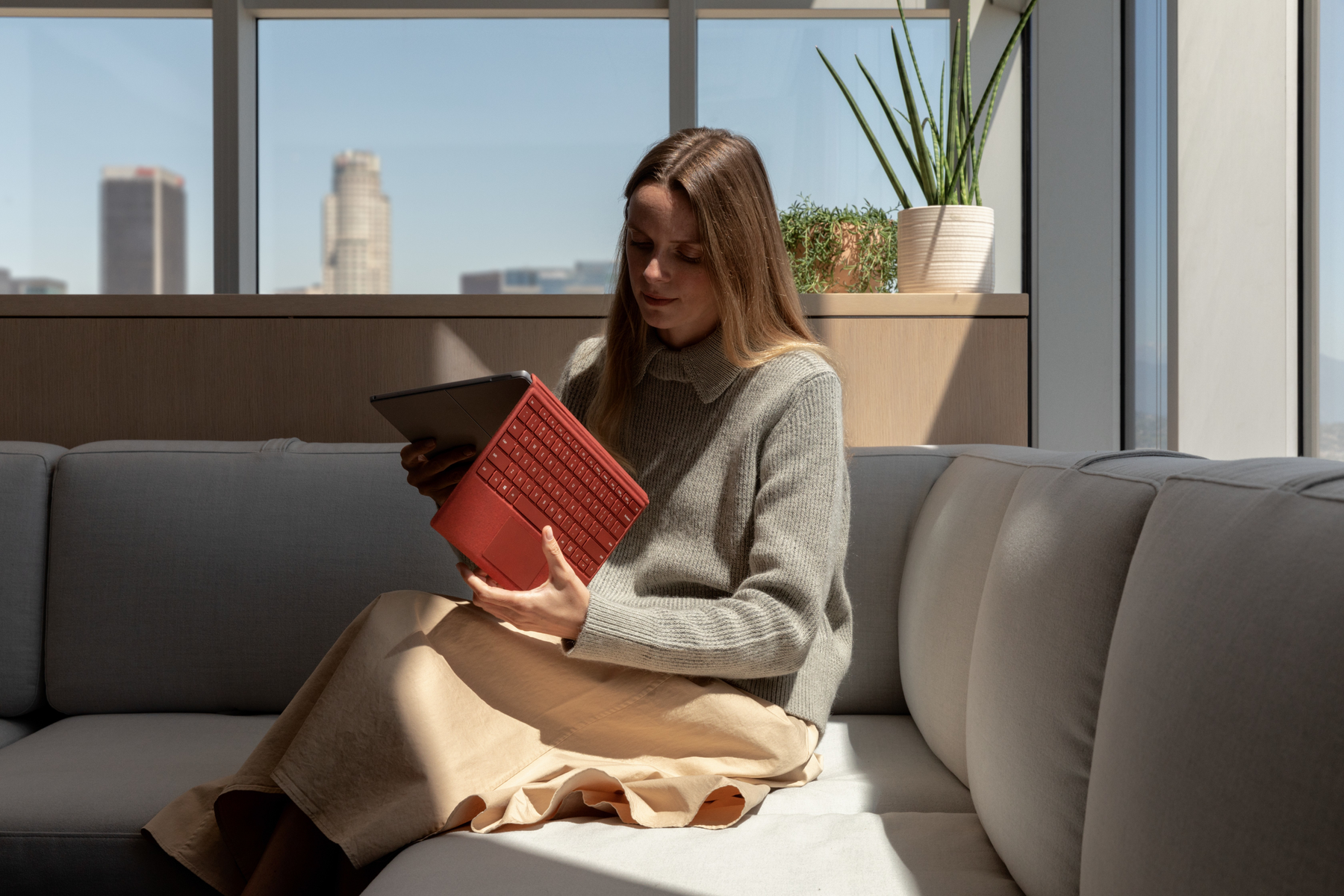 Donna seduta su un divano, tiene un tablet con tastiera rossa. Indossa un maglione grigio.