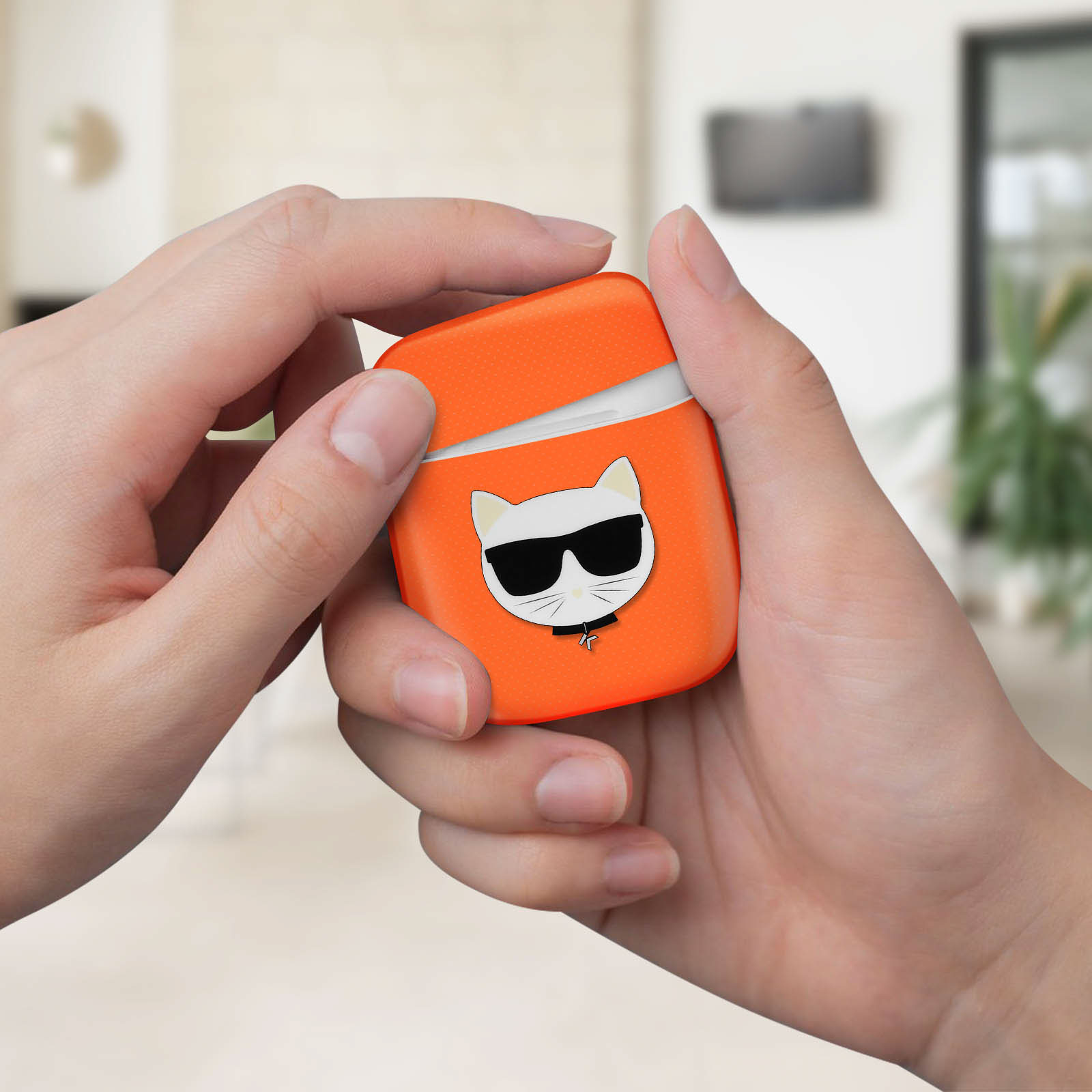 Mani che tengono una custodia arancione per AirPods con un gatto con occhiali da sole. Sfondo sfocato con finestra.