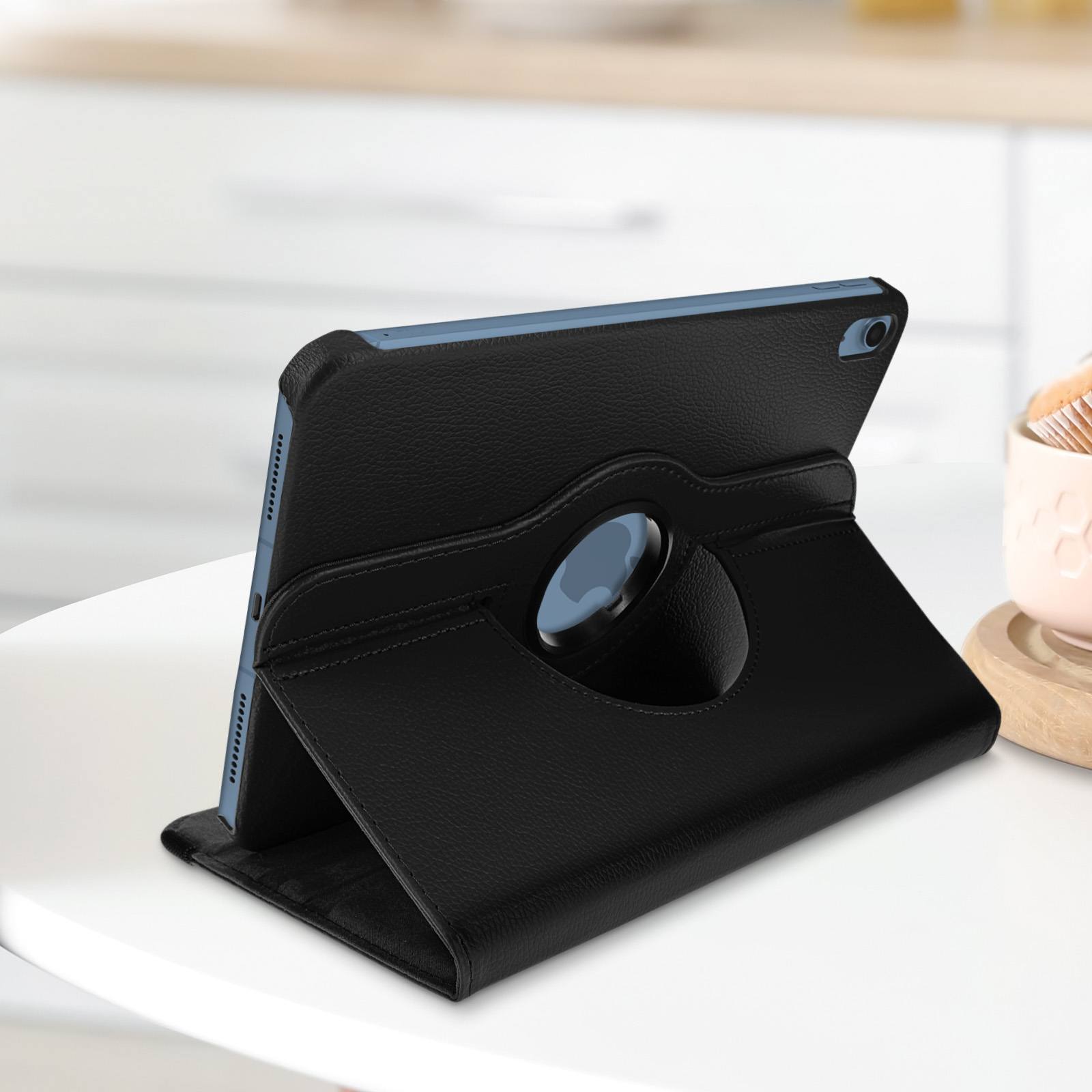 Custodia per tablet nera con supporto girevole, su un tavolo bianco. Tablet blu all'interno. Sfondo: cucina sfocata.