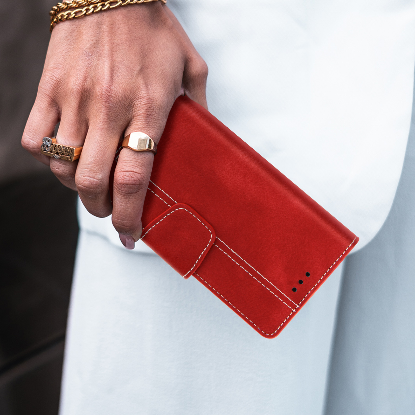 Die Hand einer Person, die eine rote Leder-Handyhülle mit weißen Nähten und Goldschmuck hält.
