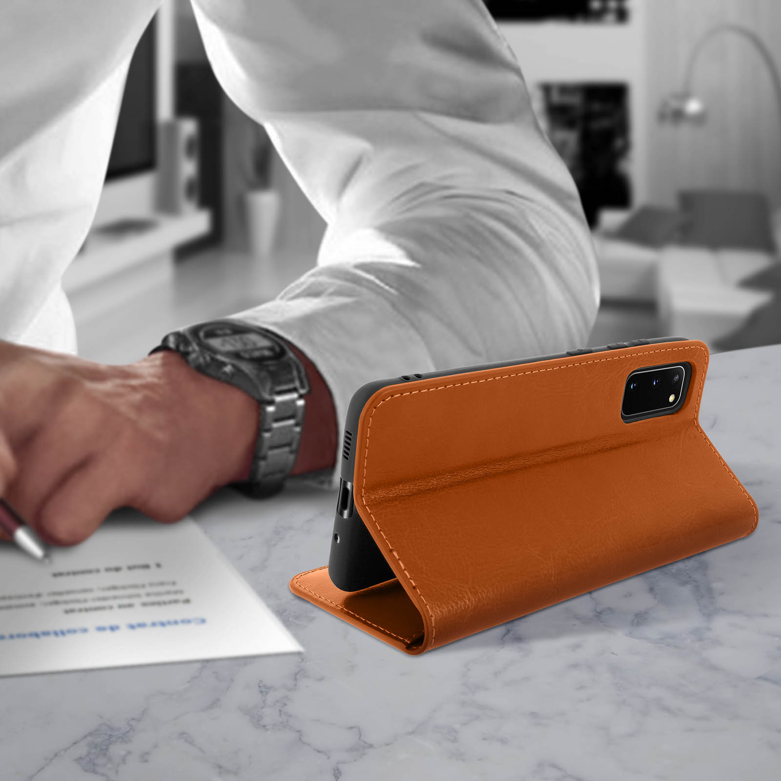 Een telefoon in een oranje leren hoesje, open op een marmeren tafel. Een persoon schrijft op de achtergrond.
