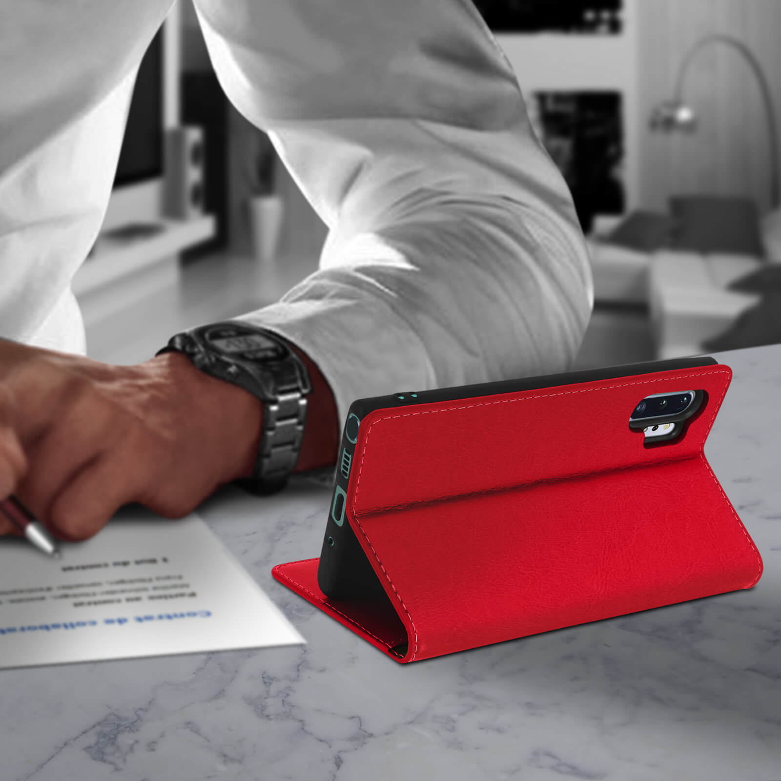Personne avec montre au poignet, stylo en main, étui rouge sur une table.