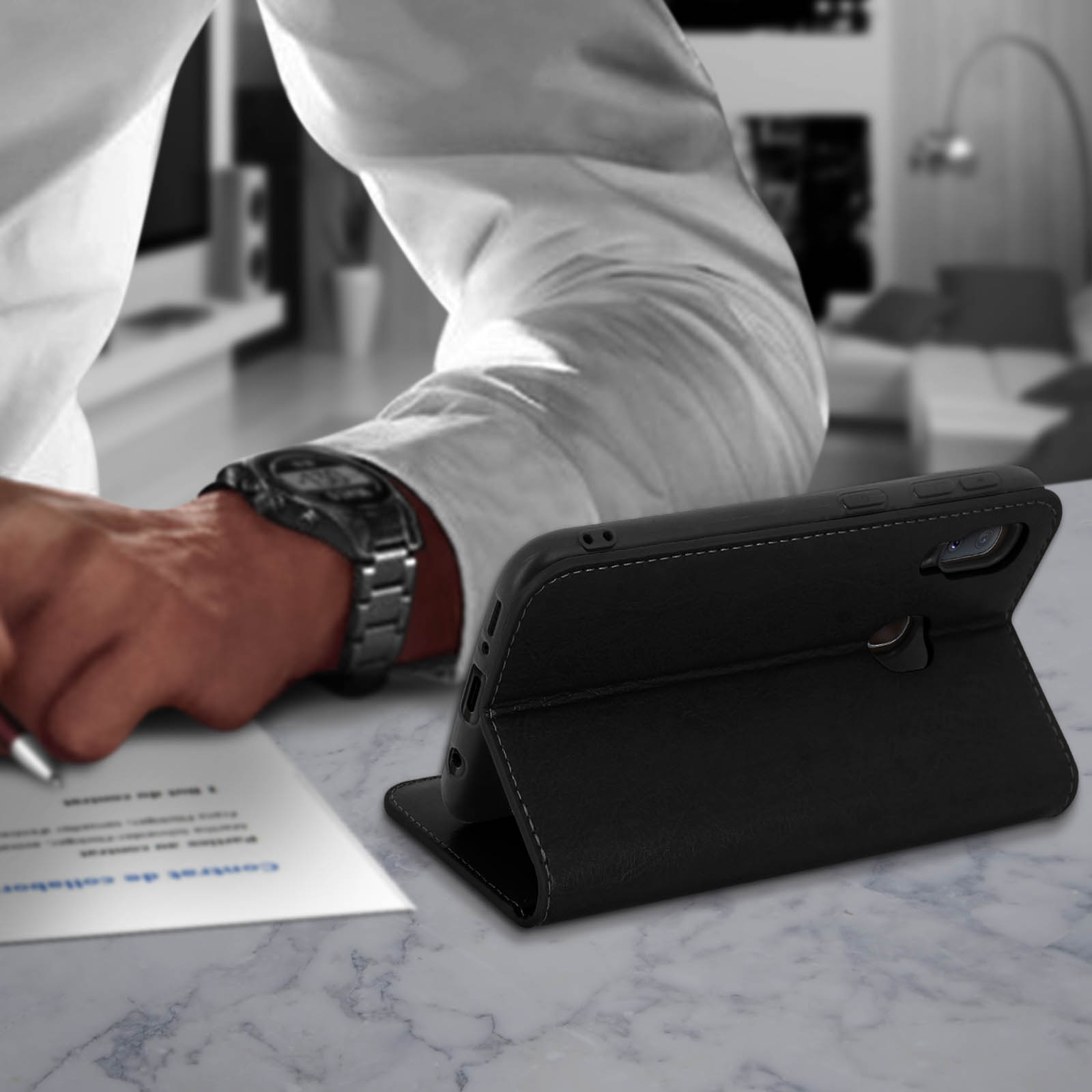 Eine schwarze Handyhülle steht offen. Die Hand eines Mannes schreibt auf Papier neben einer Uhr.