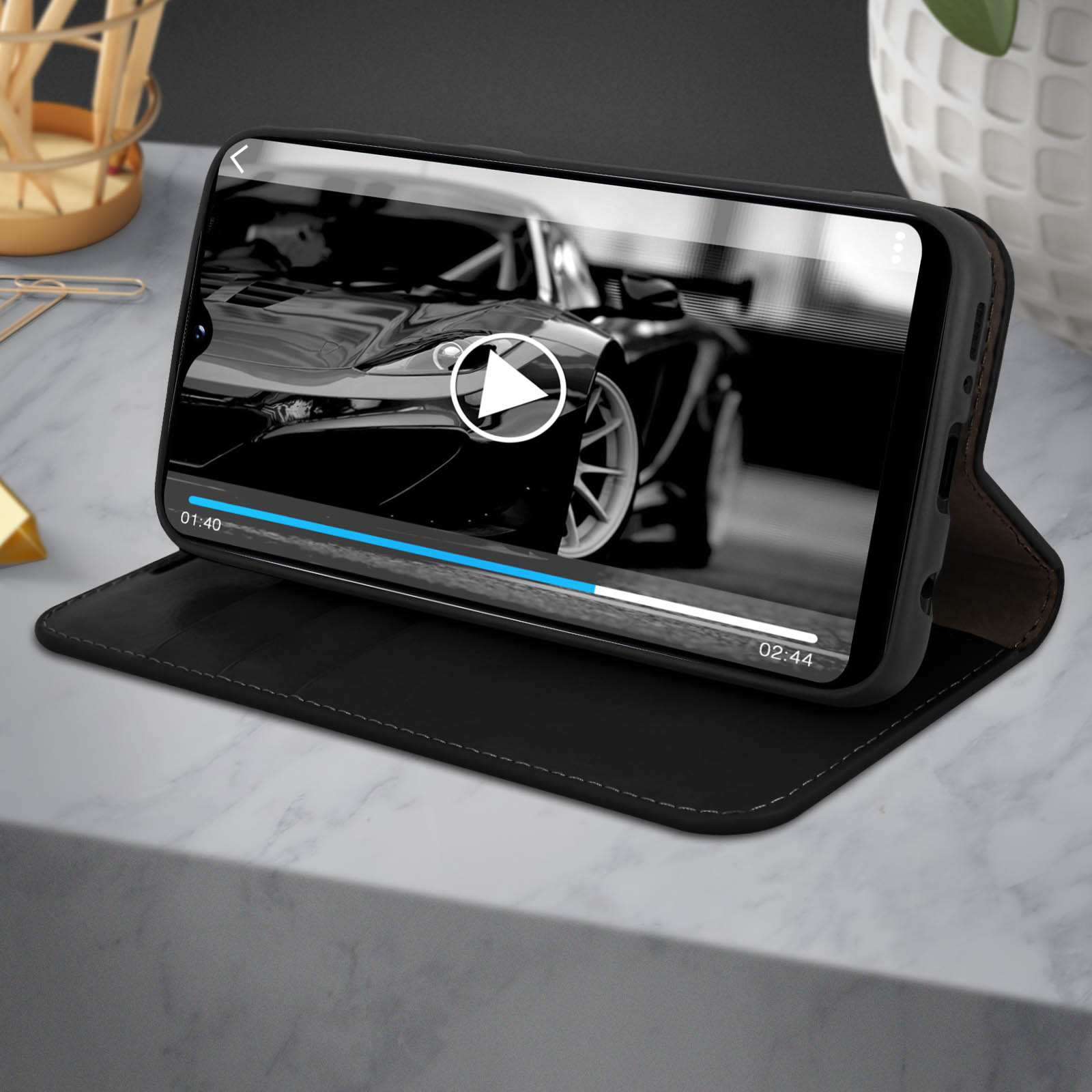Ein schwarzes Smartphone in einer Hülle, das ein Autovideo mit Wiedergabesteuerungen auf einer Oberfläche anzeigt.