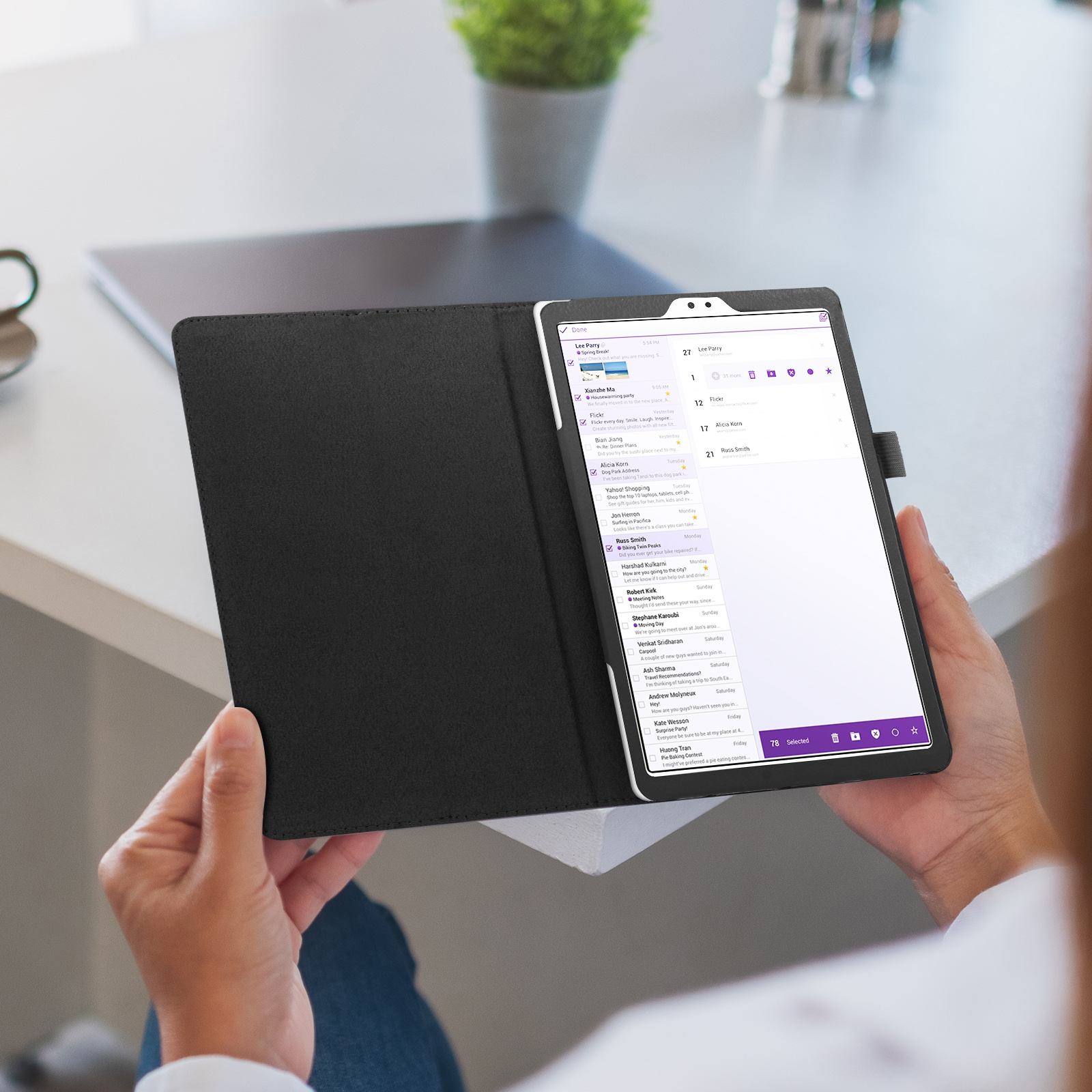 Le mani tengono una custodia nera per tablet, che mostra una casella di posta elettronica. Una pianta in vaso è sullo sfondo.