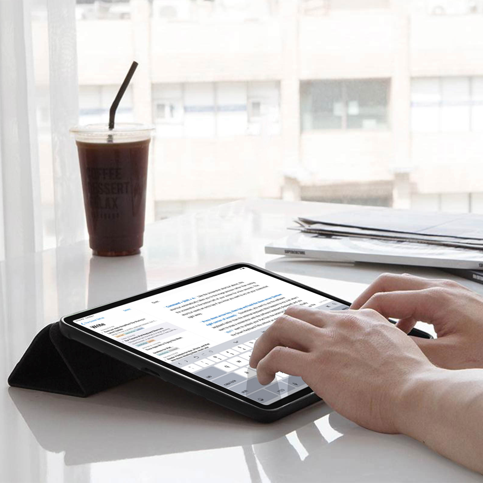 Tablet su un supporto, tastiera visibile, mani che digitano. Tazza e bevanda sul tavolo. Lo sfondo è sfocato.