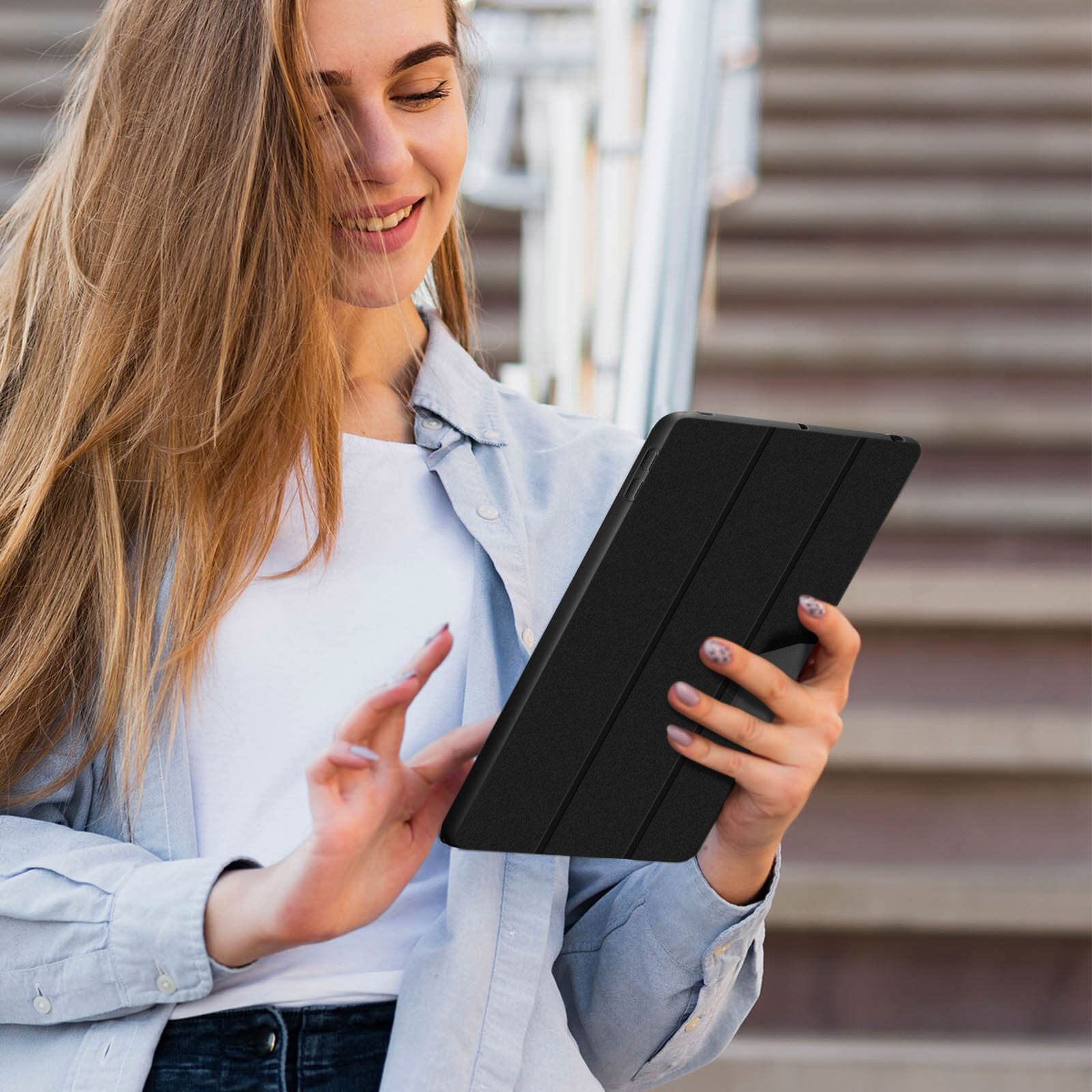 Donna che tiene un tablet nero, sorridente, all'aperto, con scale sullo sfondo.