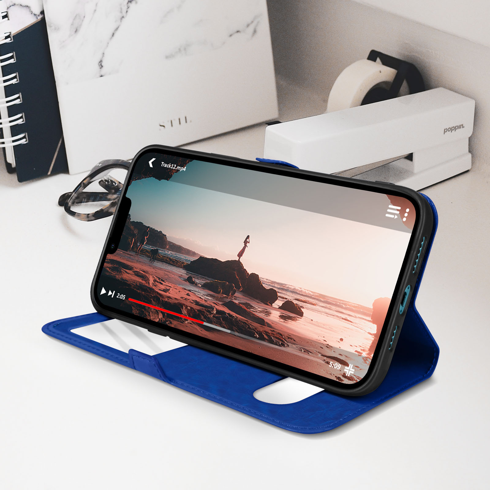 Een smartphone in een blauwe hoes toont een video op een bureau naast een bril en een nietmachine.