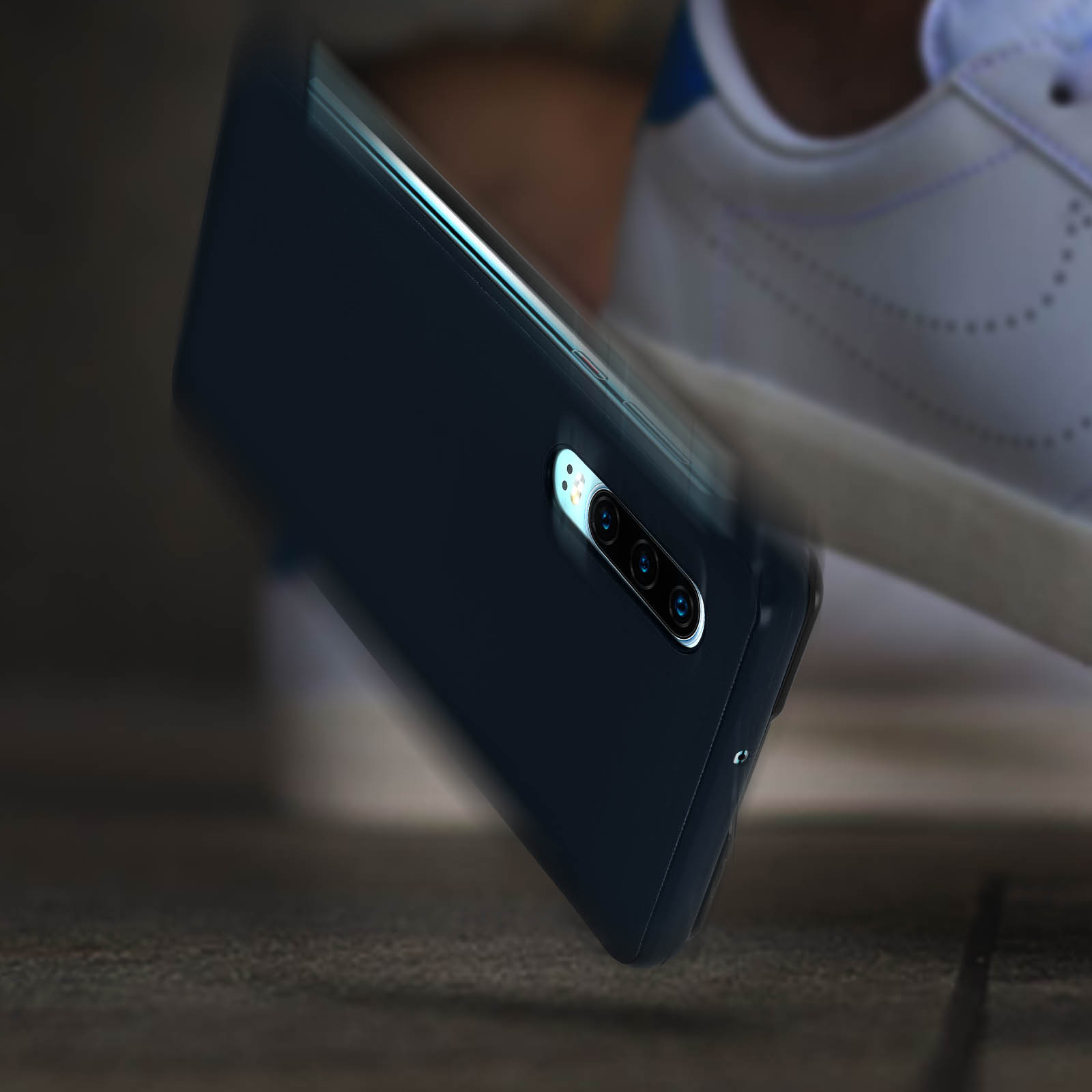 Telefono nero in custodia, angolato, cade verso una scarpa. Il telefono ha più fotocamere.