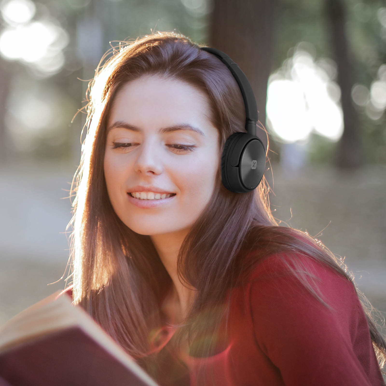 Frau mit schwarzen Kopfhörern, lächelnd, liest ein Buch in einem Park.