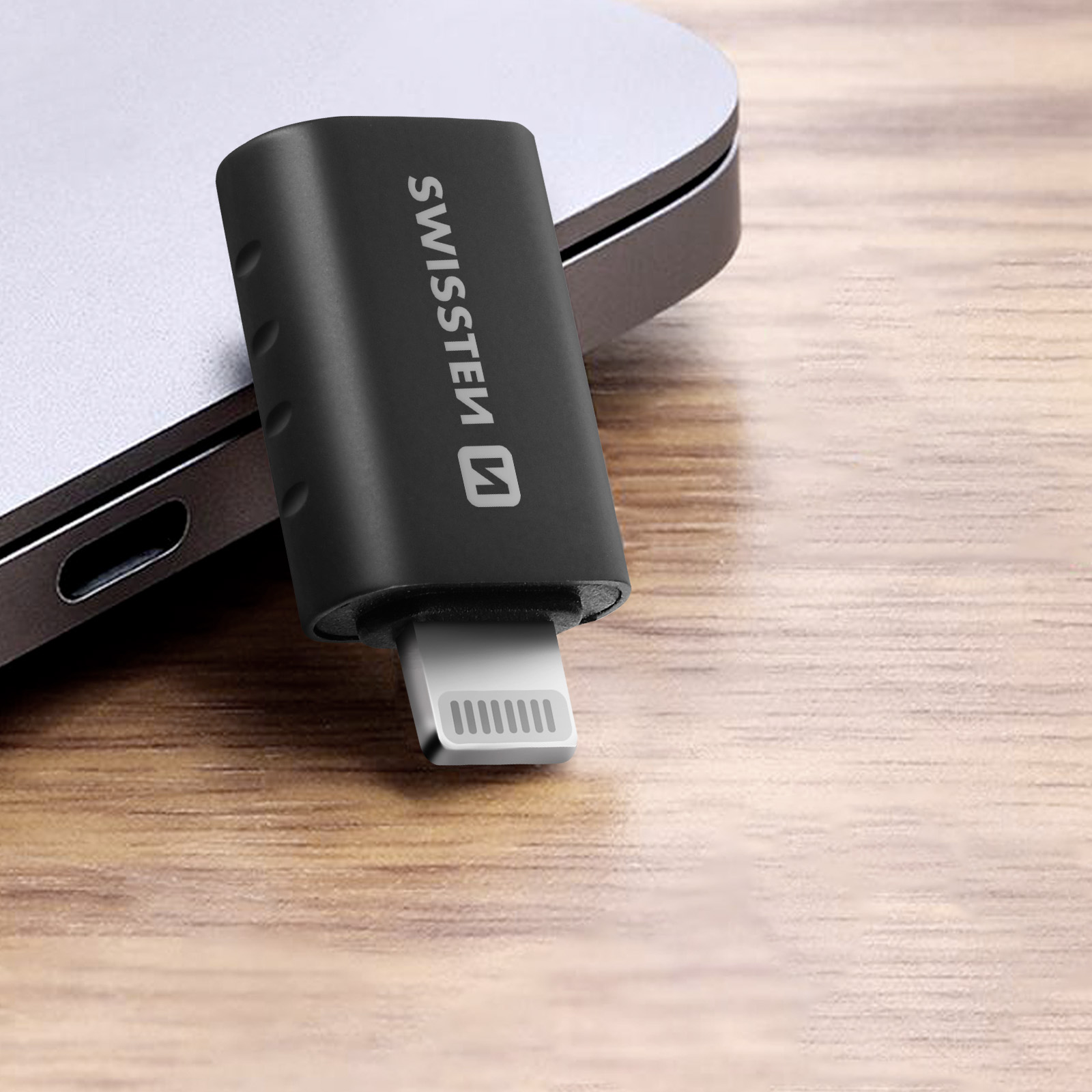 Adattatore nero da USB-C a Lightning su un tavolo di legno, accanto a un laptop.