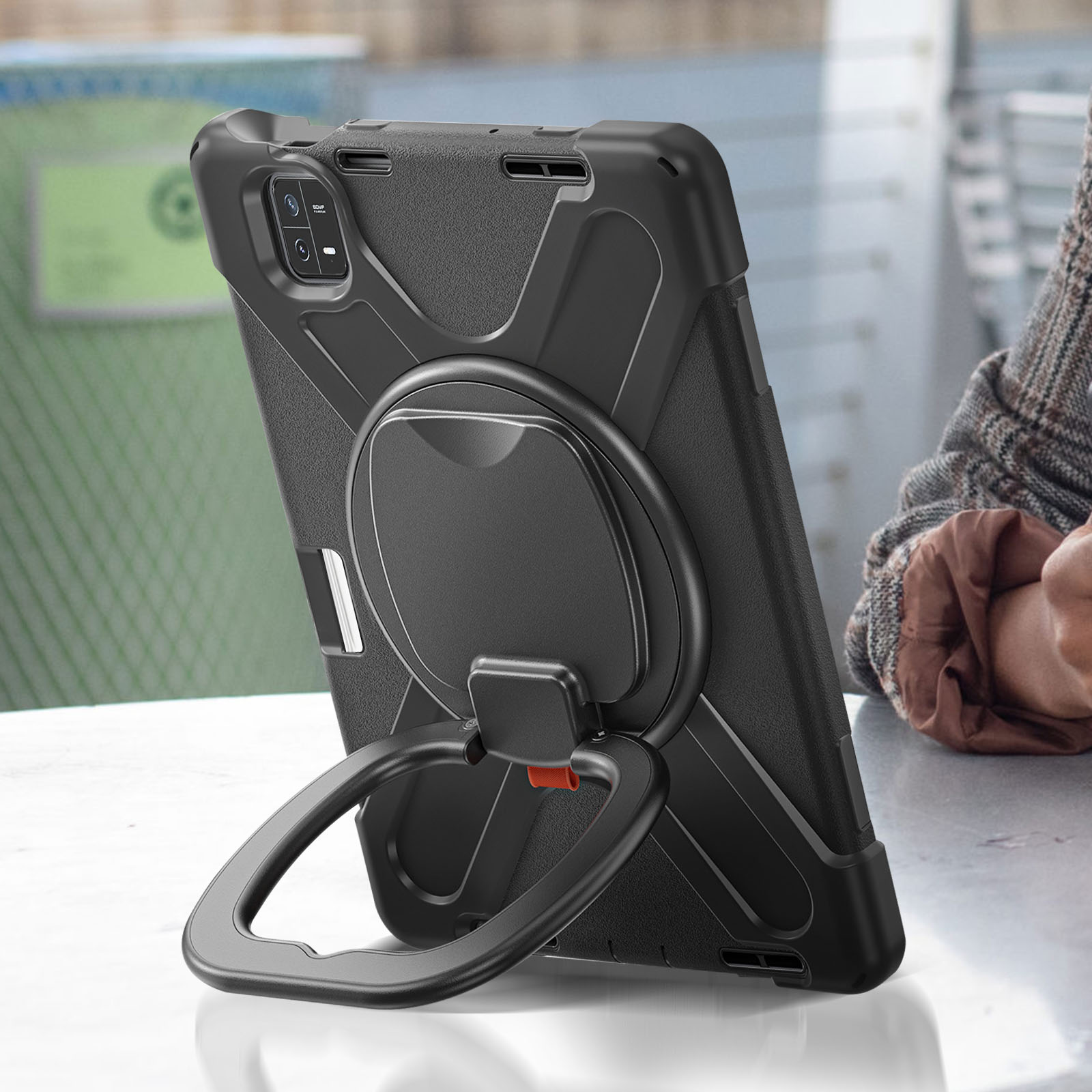 Ein schwarzes Tablet mit einem Standring steht auf einem Tisch. Eine Person in einer Jacke ist im Hintergrund.