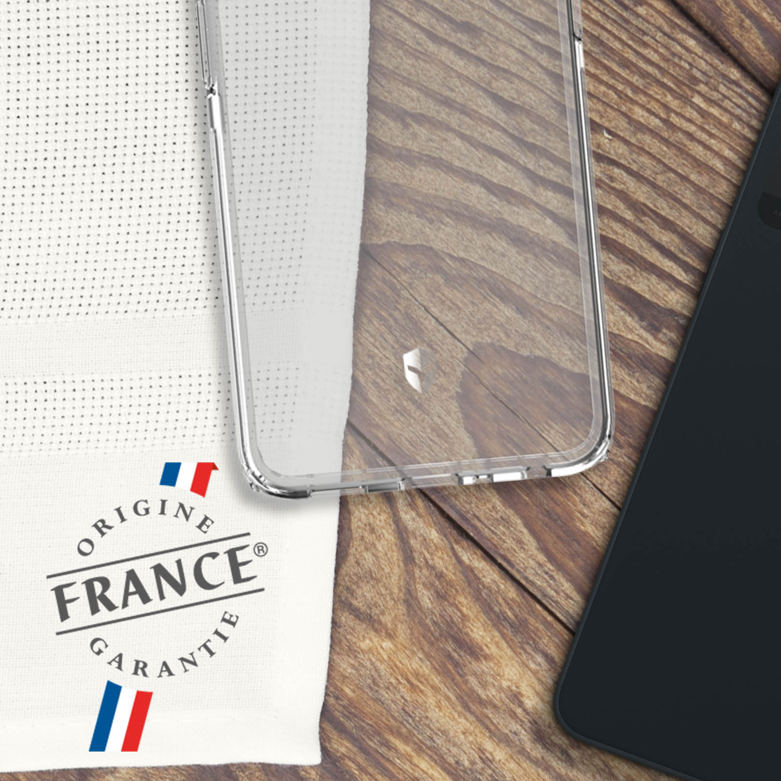 Una custodia trasparente per telefono e un panno bianco con testo 'ORIGINE FRANCE GARANTIE' su legno.