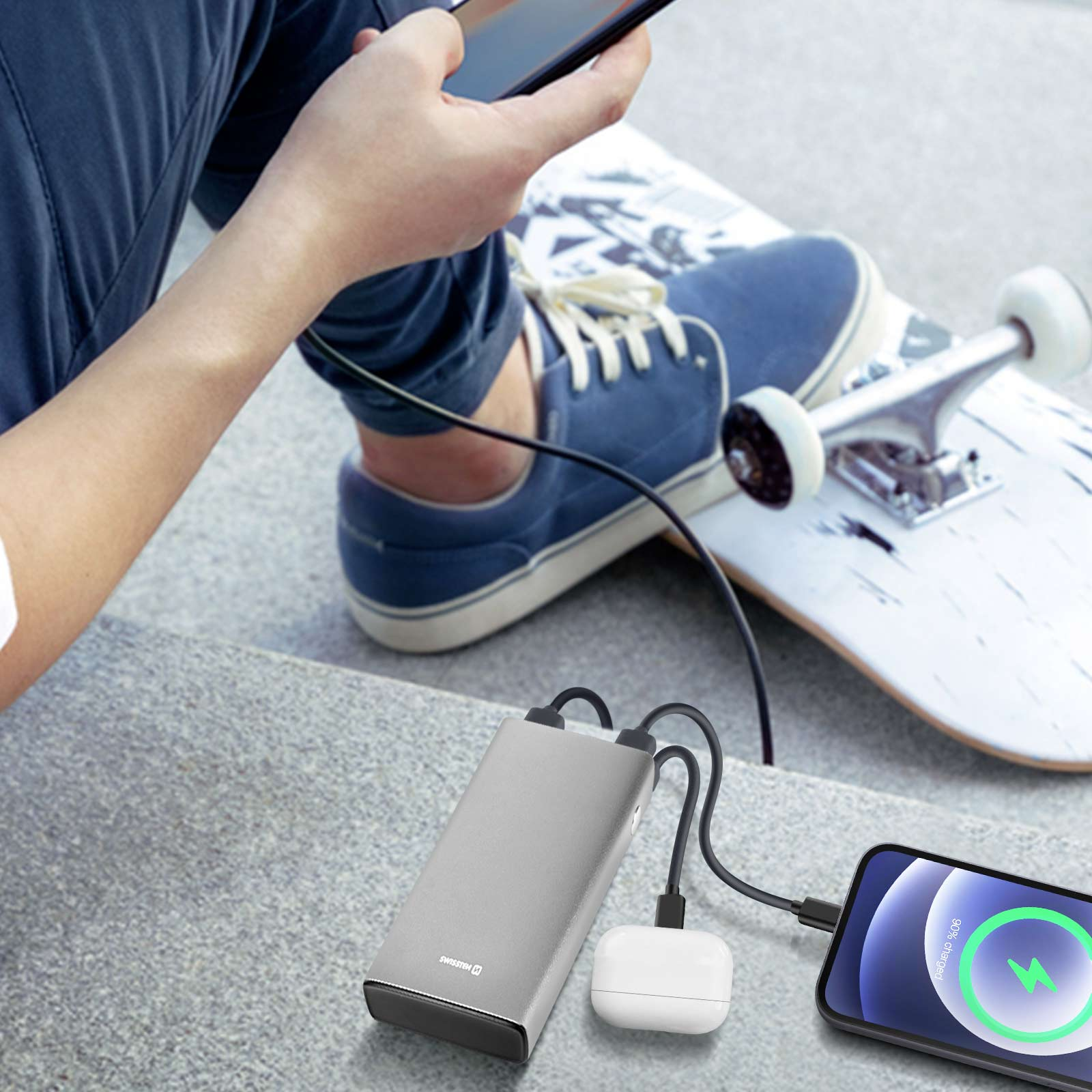 Person lädt Telefon mit Powerbank. Skateboard im Hintergrund. Graue Stufen.