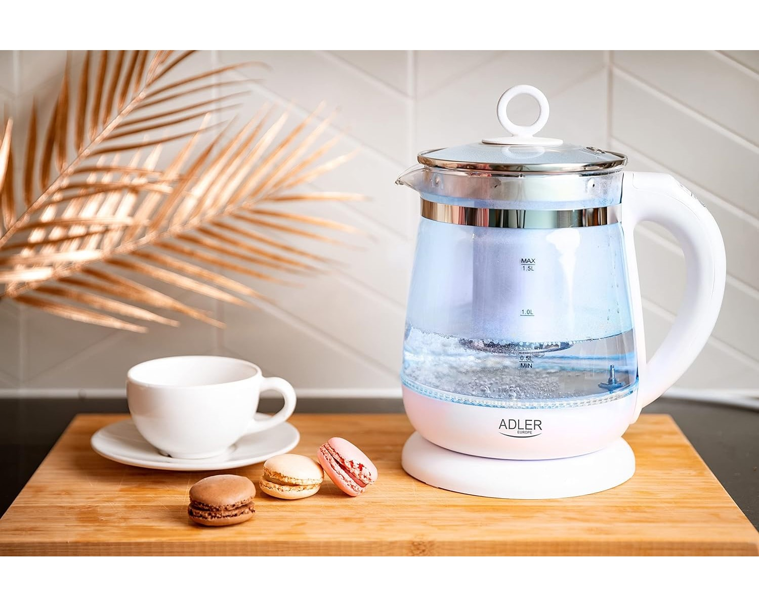 Glas-Elektrokessel mit kochendem Wasser, weißer Sockel, weiße Tasse und Makronen auf Holzbrett.