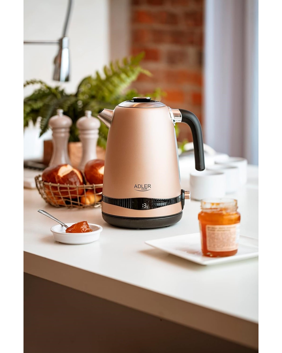 Ein roségoldener Wasserkocher mit Digitalanzeige. Brot, Marmelade und andere Gegenstände sind auf dem Tisch.