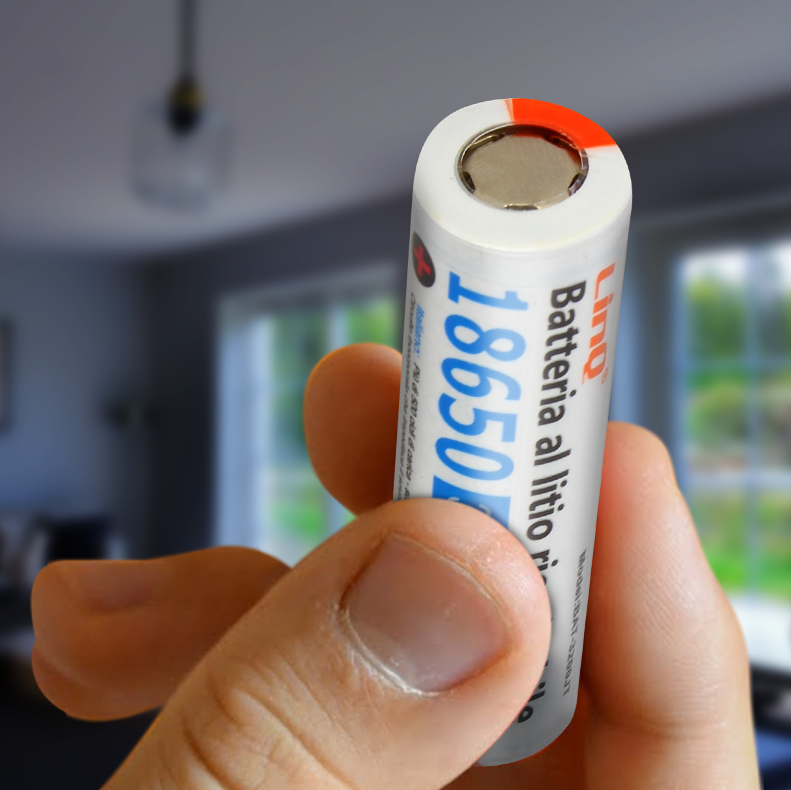 Mano che tiene una batteria cilindrica con testo, davanti a una scena interna sfocata.