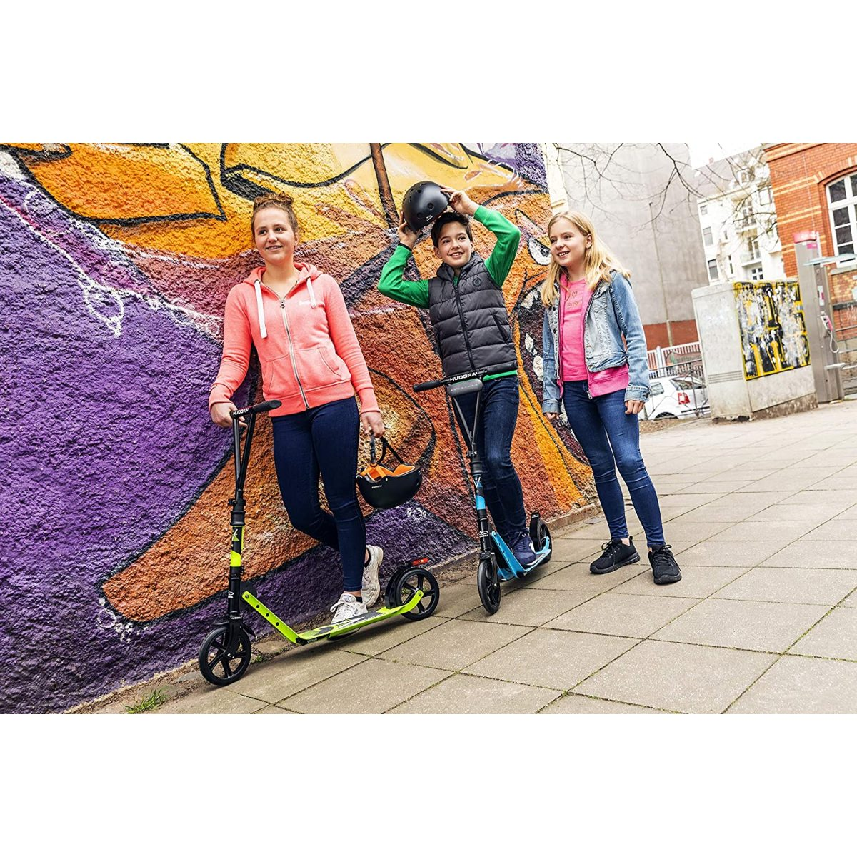 Drei Kinder mit Rollern vor einer bunten Graffiti-Wand.