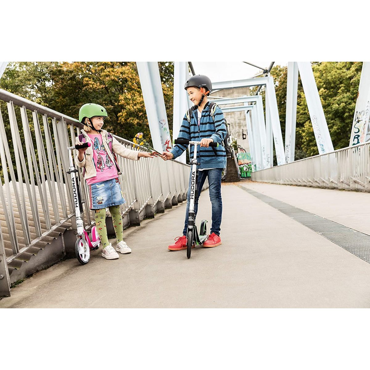 Zwei Kinder auf Rollern auf einer Brücke. Sie tragen Helme. Eines hält Blumen.