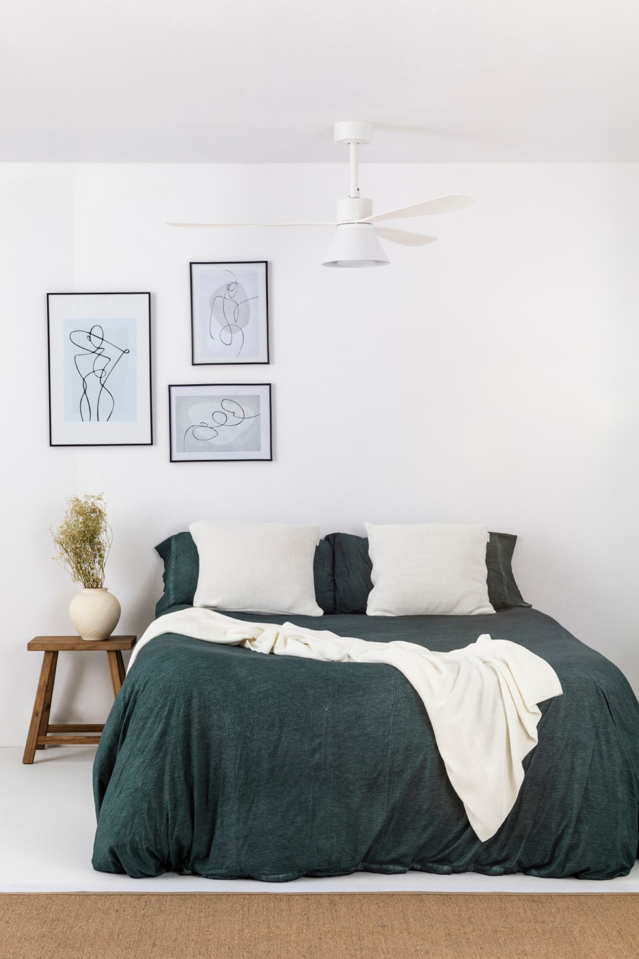 Schlafzimmer mit dunkelgrünem Bett, weißer Bettwäsche, Kunstwerken, kleinem Holztisch, Deckenventilator.