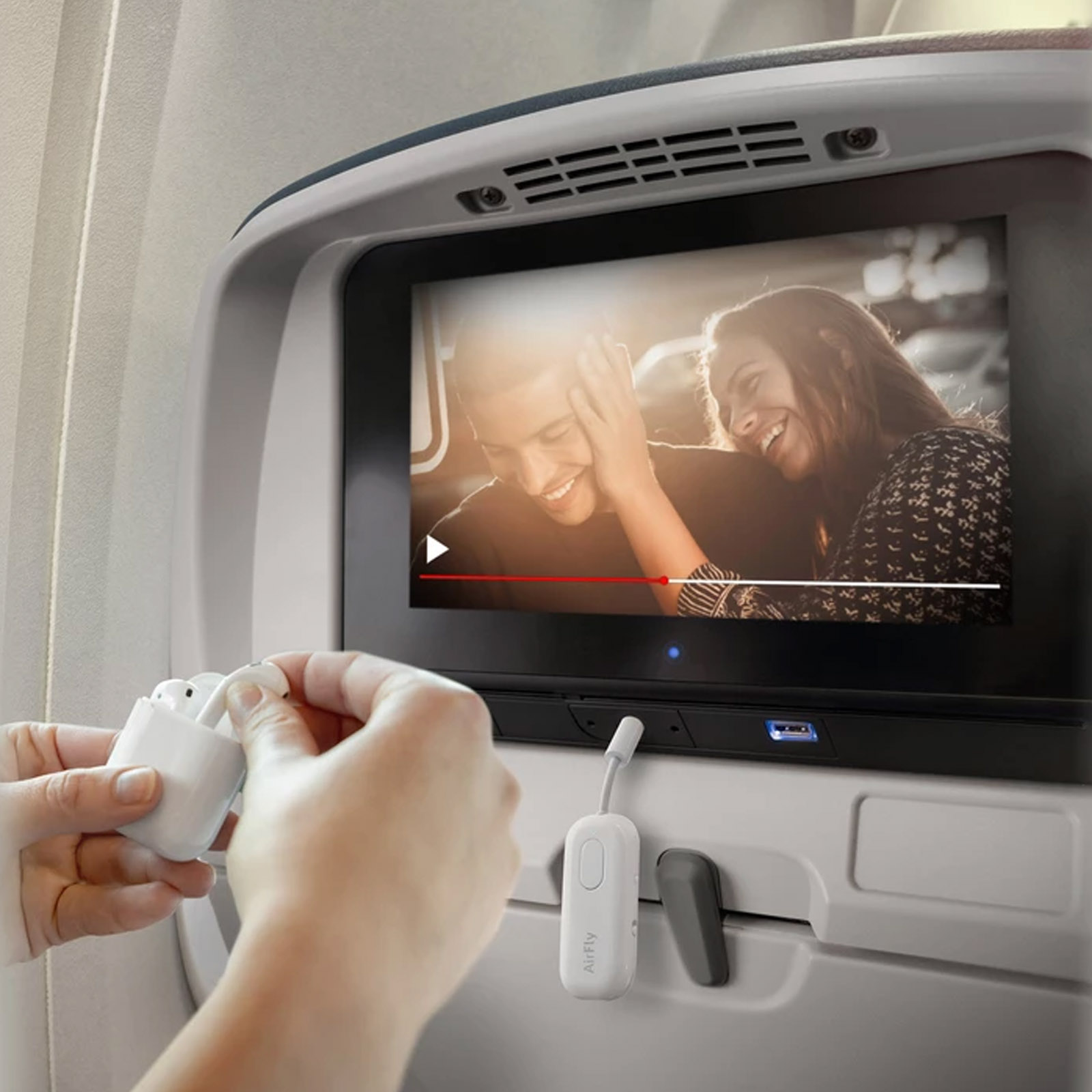 Eine Person hält Airpods in der Nähe eines Bildschirms, auf dem ein Paar in einem Flugzeug ein Video anschaut.