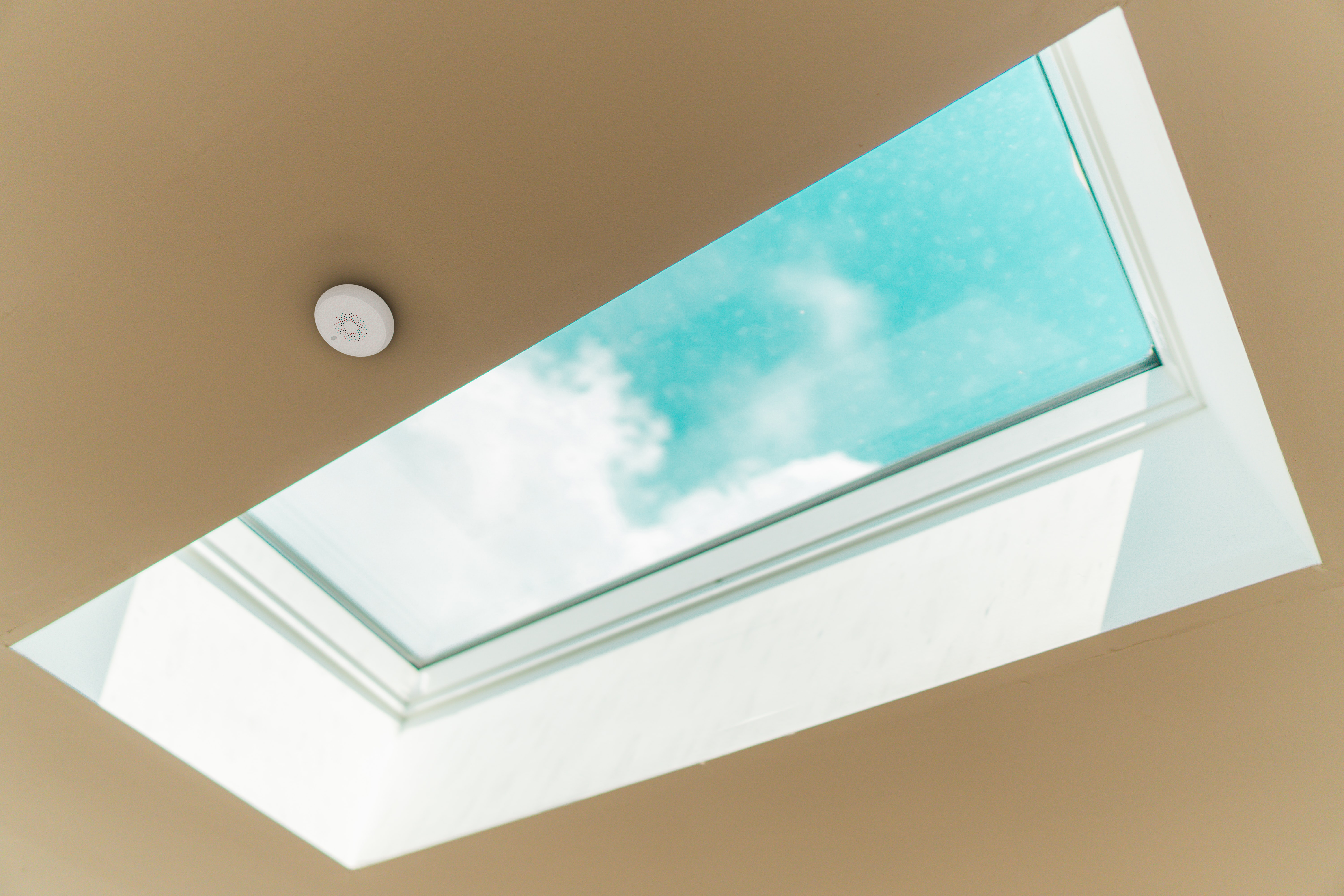Blick von unten auf ein Oberlicht. Es ist ein rechteckiges Fenster in einer beigen Decke, das einen blauen Himmel mit Wolken zeigt.