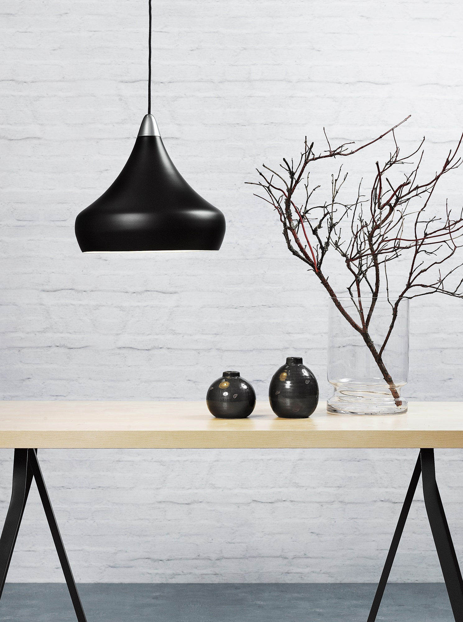 Eine schwarze Pendelleuchte hängt über einem Holztisch. Zwei kleine Vasen und eine große Vase mit Ästen stehen auf dem Tisch.