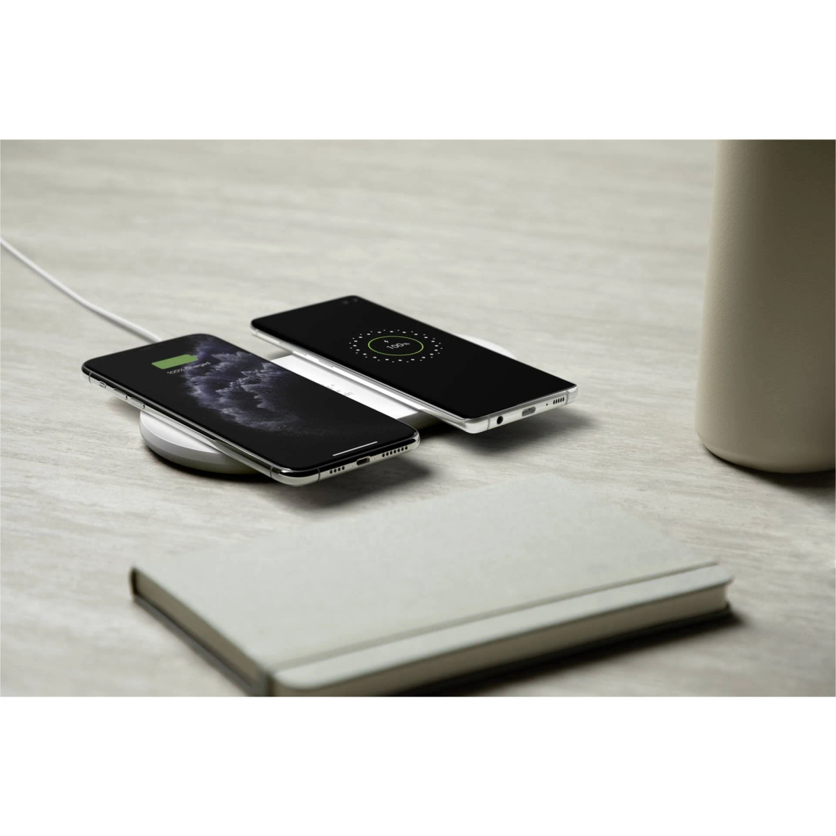 Zwei Smartphones laden auf einem weißen kabellosen Ladegerät, daneben ein Notizbuch auf einer hellen Oberfläche.