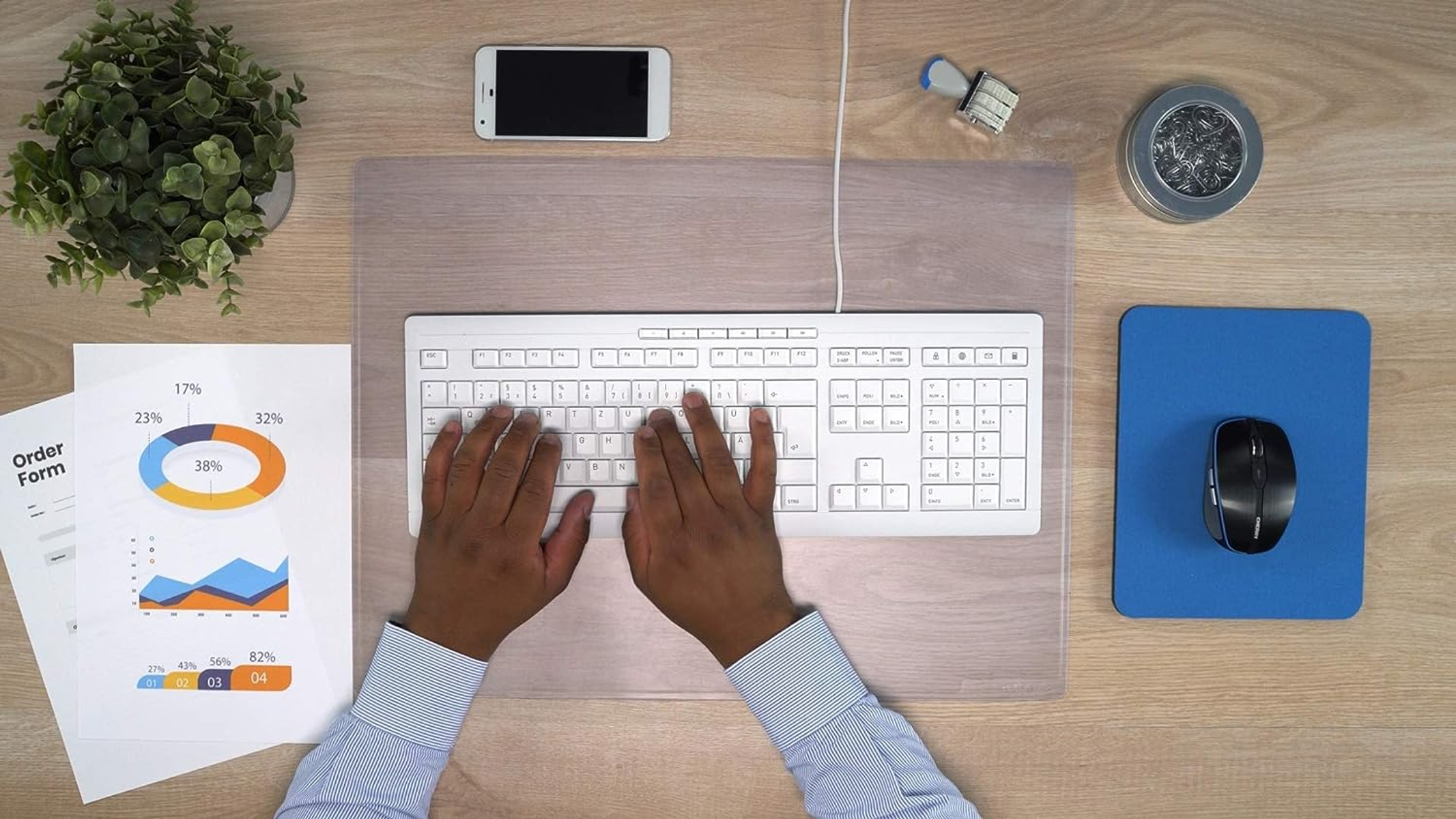 Widok z góry na biurko: klawiatura, mysz, papiery z wykresami i smartfon. Drewniana powierzchnia.