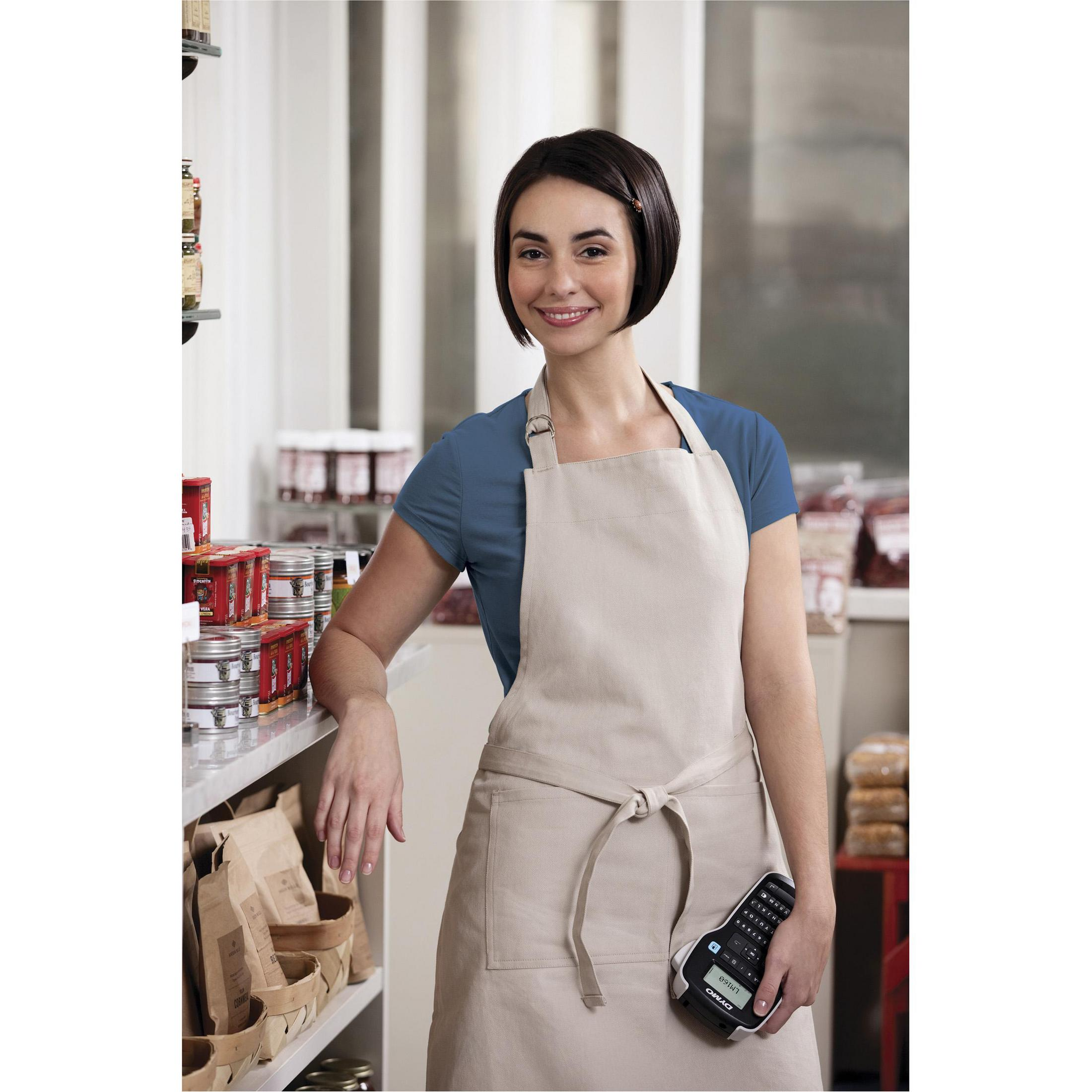 Femme en tablier sourit, tenant une étiqueteuse dans une boutique. Étagères avec produits en arrière-plan.