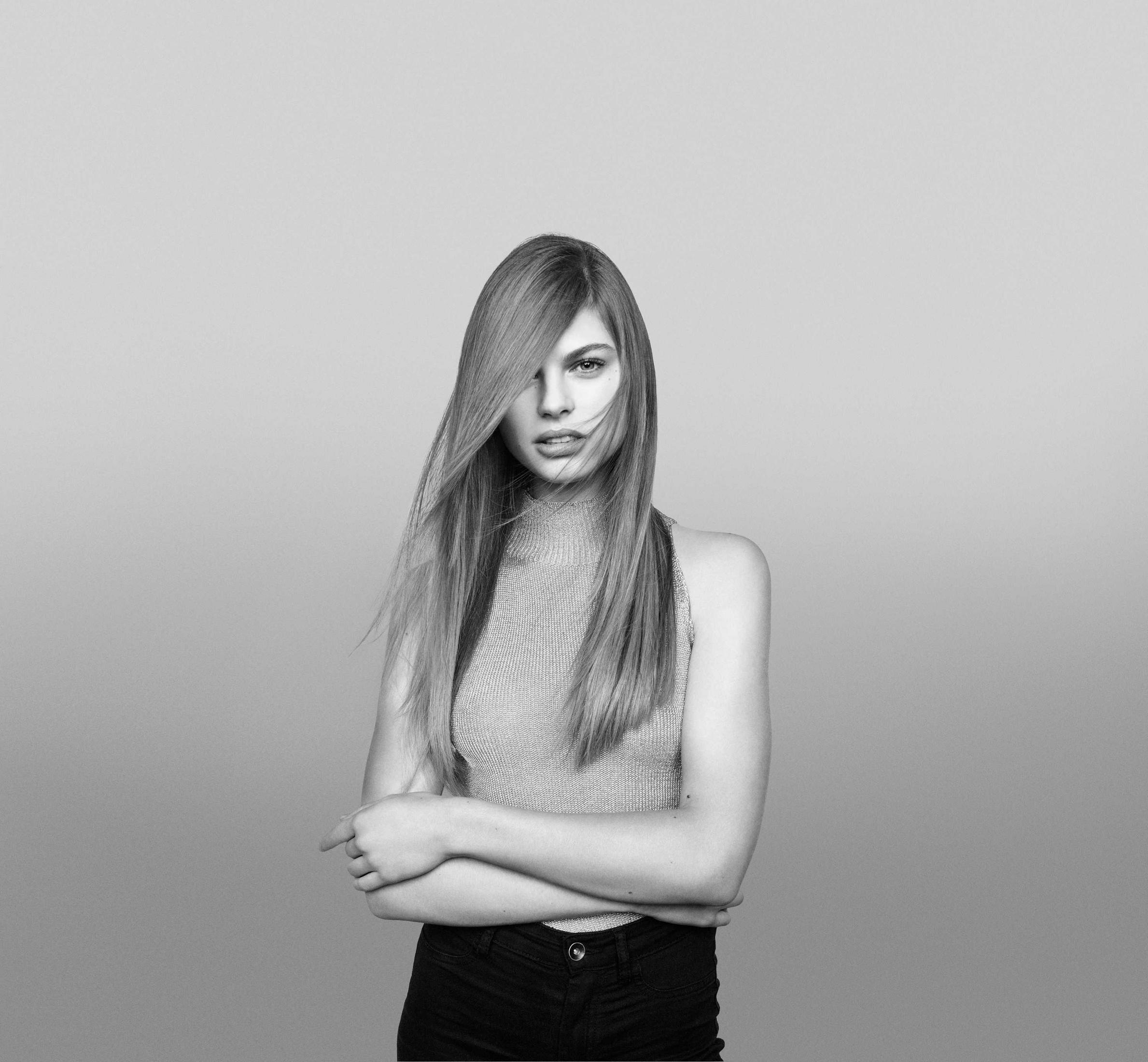 Kobieta z długimi włosami i jasnym topem patrzy w kamerę. Ma na sobie ciemne spodnie.