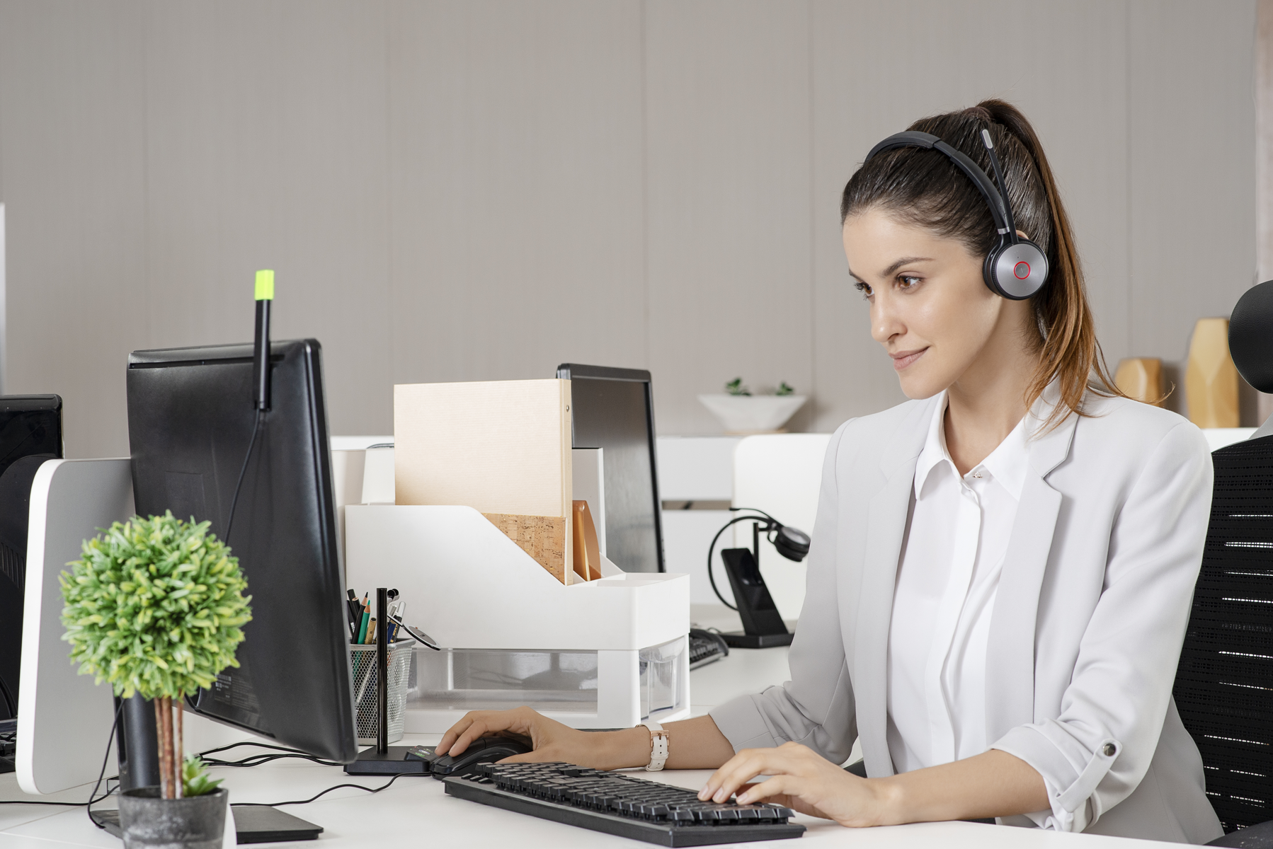 Eine Frau in grauem Anzug mit Headset, die im Büro am Computer arbeitet.