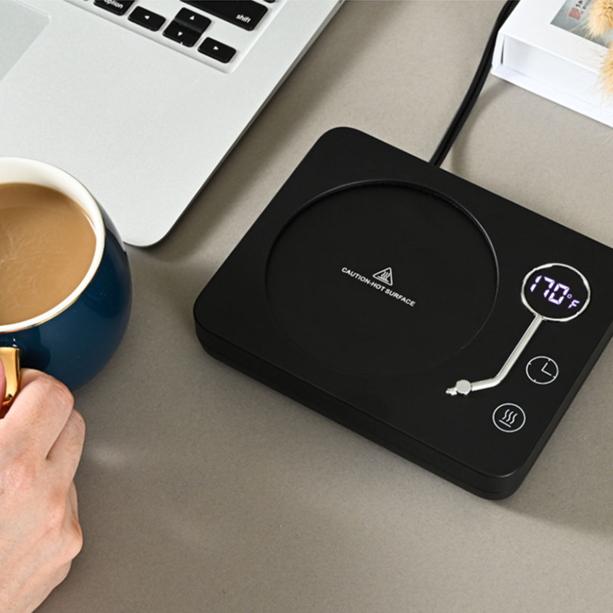 Schwarzer Kaffee-Wärmer mit Digitalanzeige, Kaffeetasse und Laptop im Hintergrund auf grauer Oberfläche.