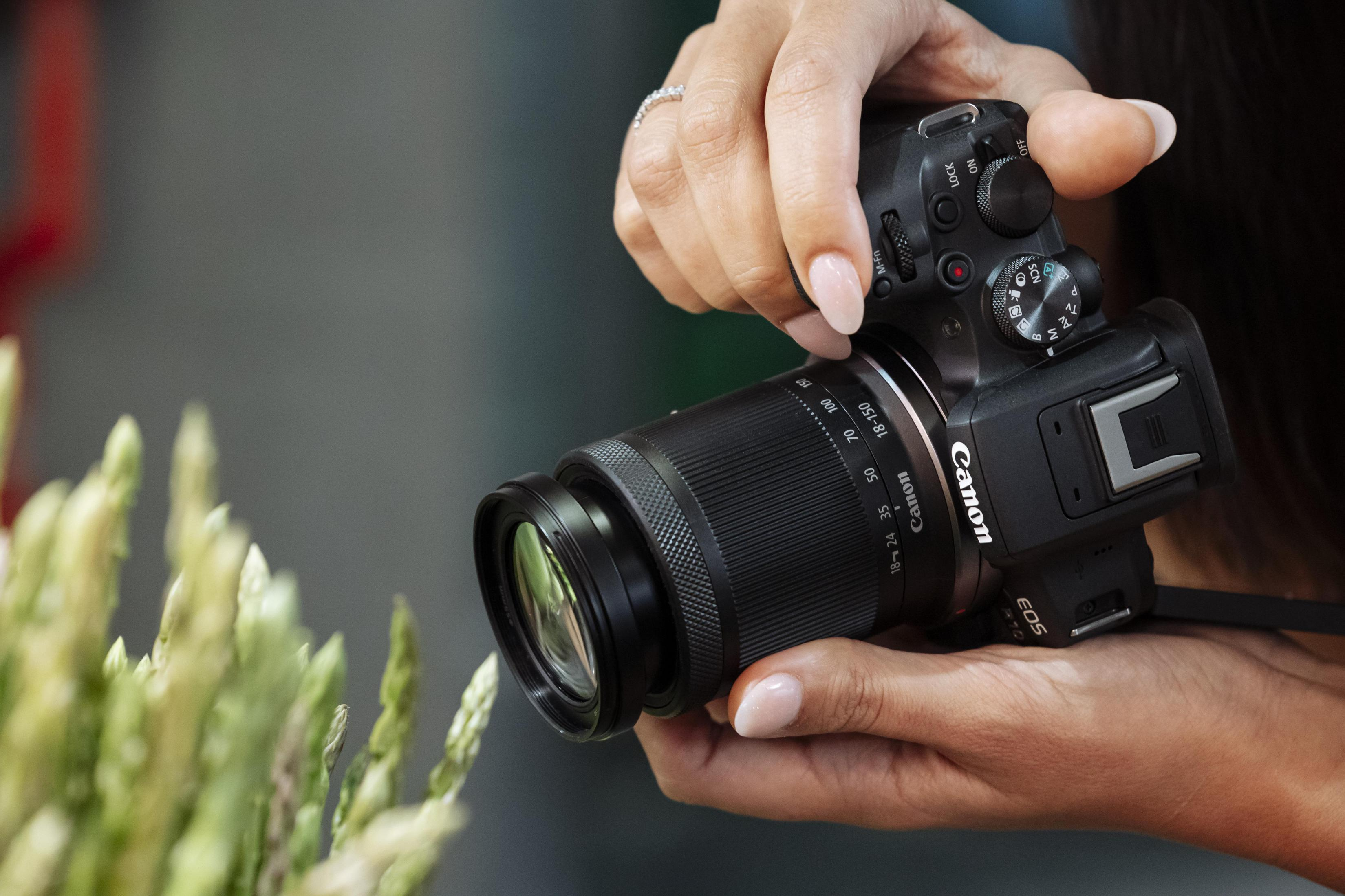 Osoba trzymająca czarny aparat Canon z obiektywem, skupiająca się na zielonych roślinach.