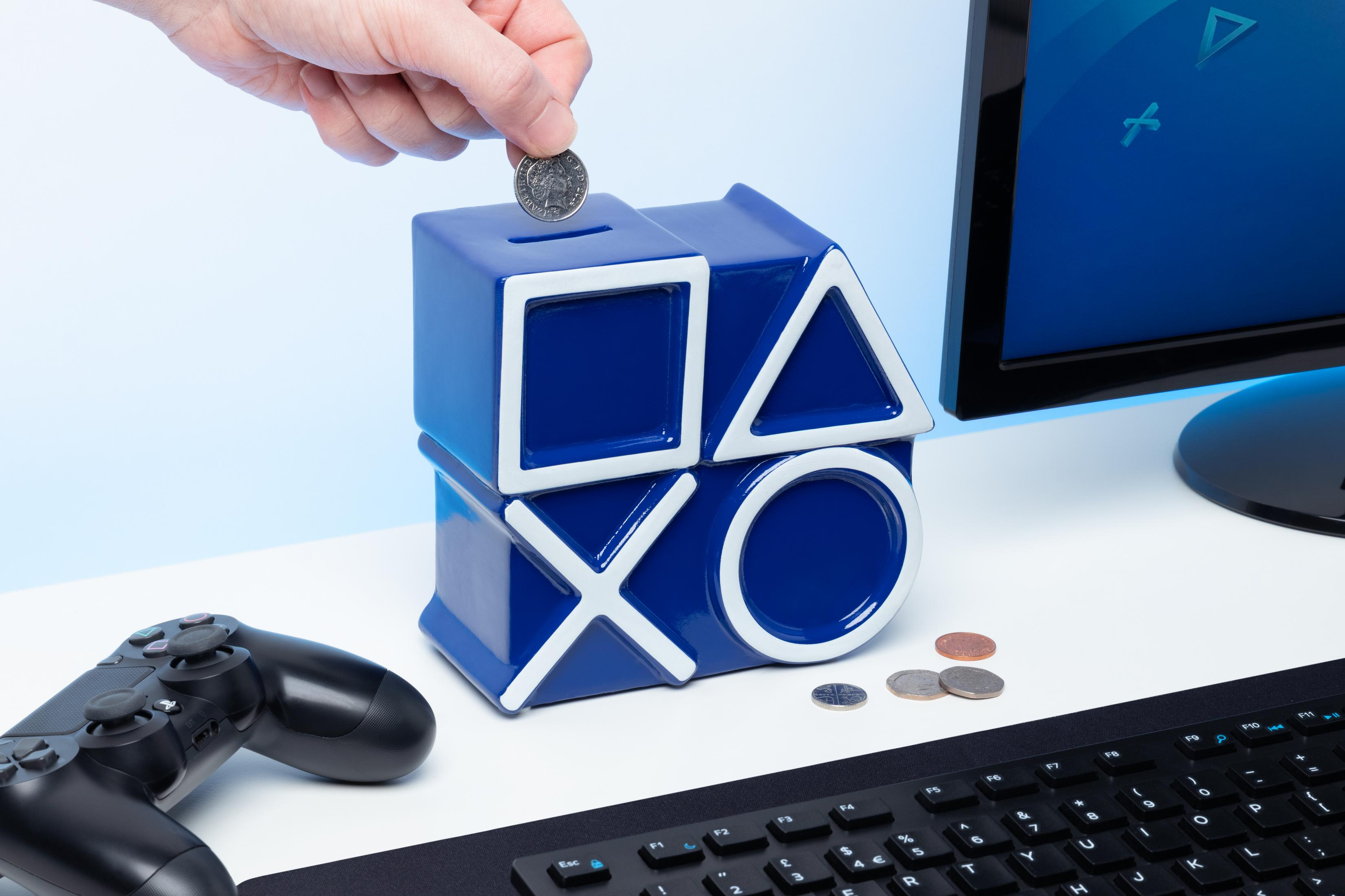Hand wirft Münze in eine blaue PlayStation-Spardose. Andere Münzen, Controller und Monitor sind sichtbar.